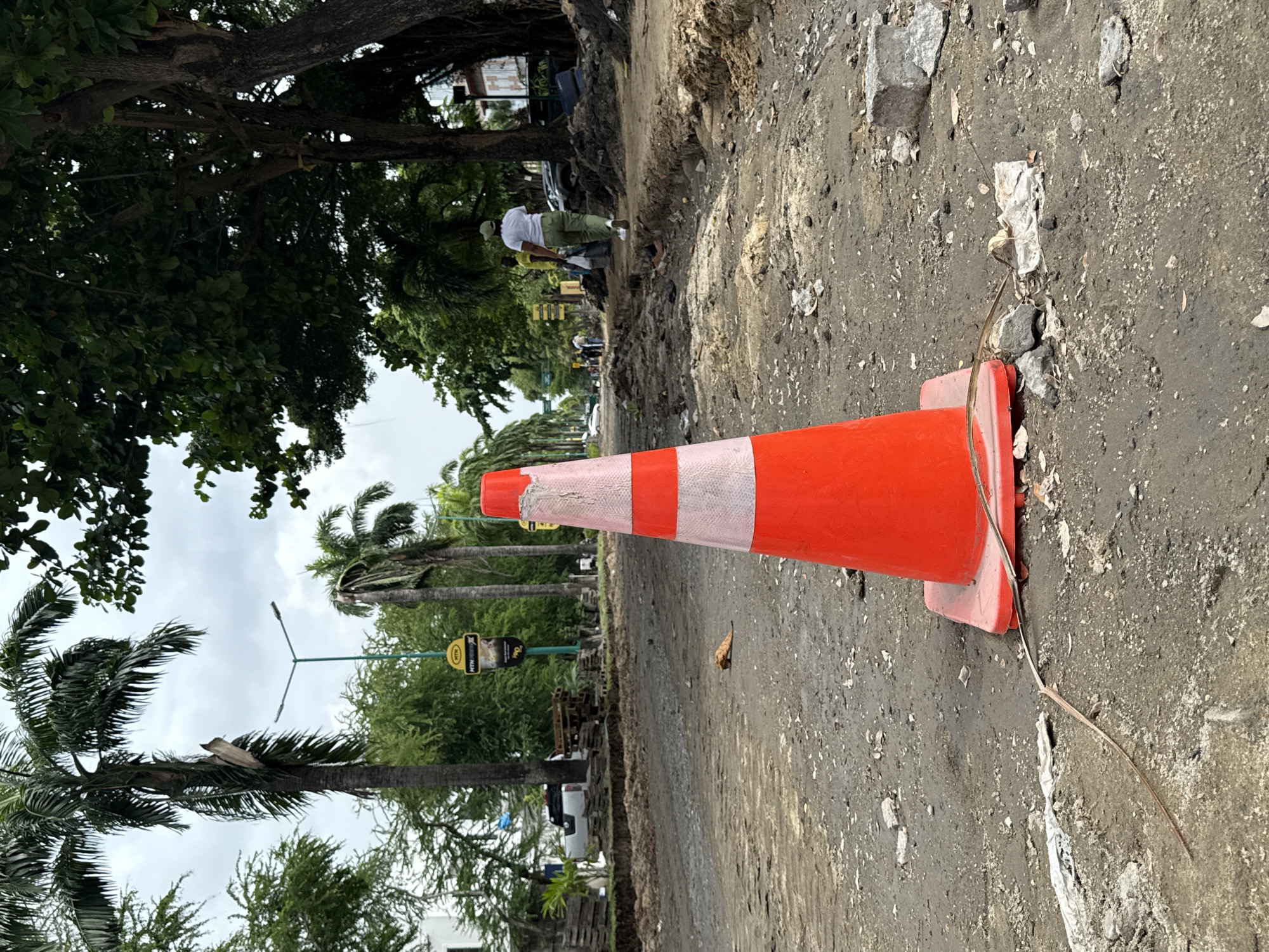 An eye-level outdoor shot reveals a construction site in Lagos, Nigeria, signified by a large orange and white traffic cone lying on its side in the foreground. The road is heavily eroded, and debris is scattered across the asphalt. In the background, a construction crew works on the road under the shade of trees. Palm trees and lush greenery indicate a tropical environment. Utility poles and street signs are present, along with parked vehicles. The sky is overcast.