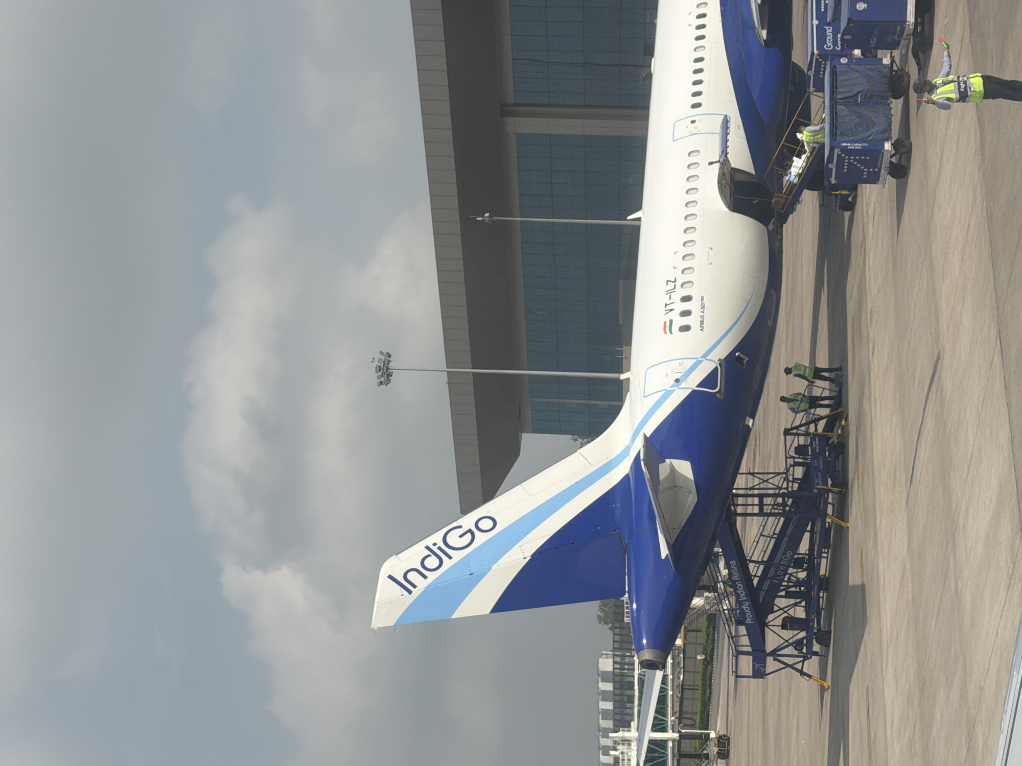 An Indigo Airlines airplane, registered as VT-ILZ and an Airbus A321neo, is parked on the tarmac at an airport. The aircraft's tail, adorned with the Indigo logo, is visible against a cloudy sky. A jet bridge or airstair is connected to the airplane, and several ground crew members are present. Some individuals are near the aircraft and the airstair, and one person in a high-visibility vest is directing traffic with illuminated batons. Two blue baggage carts are positioned near the aircraft. In the background, a modern airport terminal building with large, reflective windows and a gray overhang is visible. The scene is likely at an airport near Vikram Nagar, India, given the presence of Indigo Airlines, an Indian carrier.