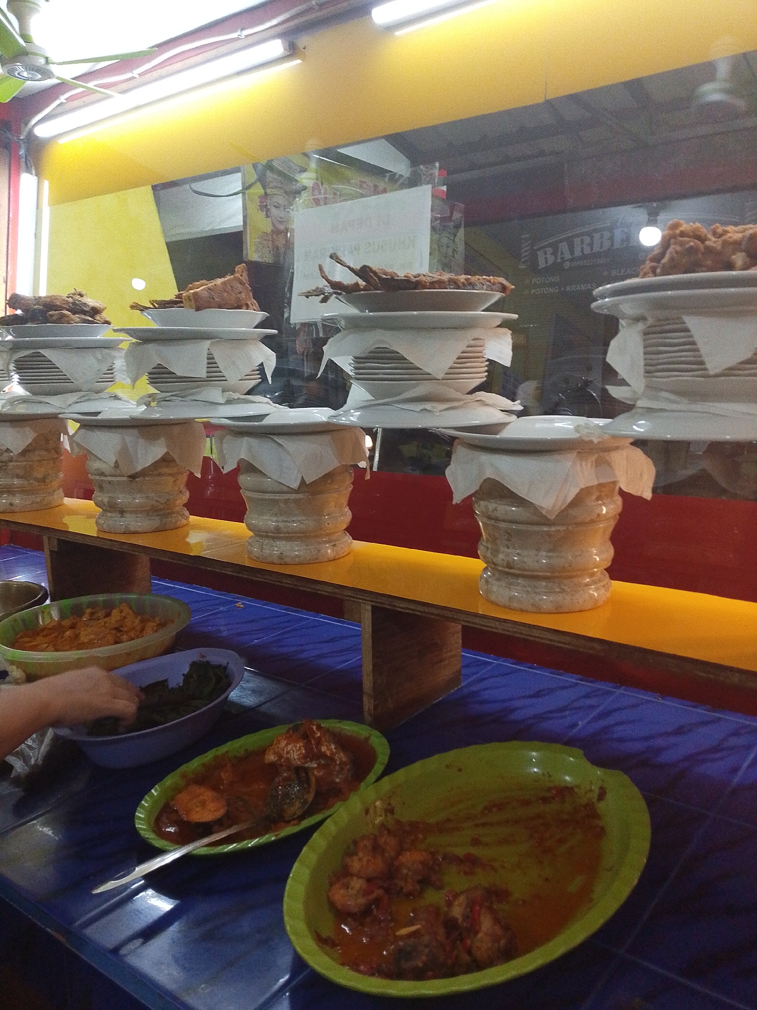 The image depicts a food stall, likely in Kediri City, Indonesia, displaying a variety of dishes. In the foreground, two oval green plates hold food. One plate contains pieces of meat in a red sauce, with a spoon resting on it. The other plate has less food, primarily sauce and some meat remnants. Behind these, a person's hand is visible reaching into a purple bowl filled with dark green leafy vegetables. To the left of the purple bowl is a large green bowl containing an orange-colored dish with visible chunks of food.
In the background, a yellow shelf displays stacked white plates, with some plates holding portions of fried or grilled meat dishes. These stacked plates are supported by decorative, cylindrical bases. A reflection in the glass behind the food stall shows a ceiling fan, fluorescent lights, and signage that appears to be for a barber shop, with text like "BARBER" visible. The overall environment suggests a casual dining setting.