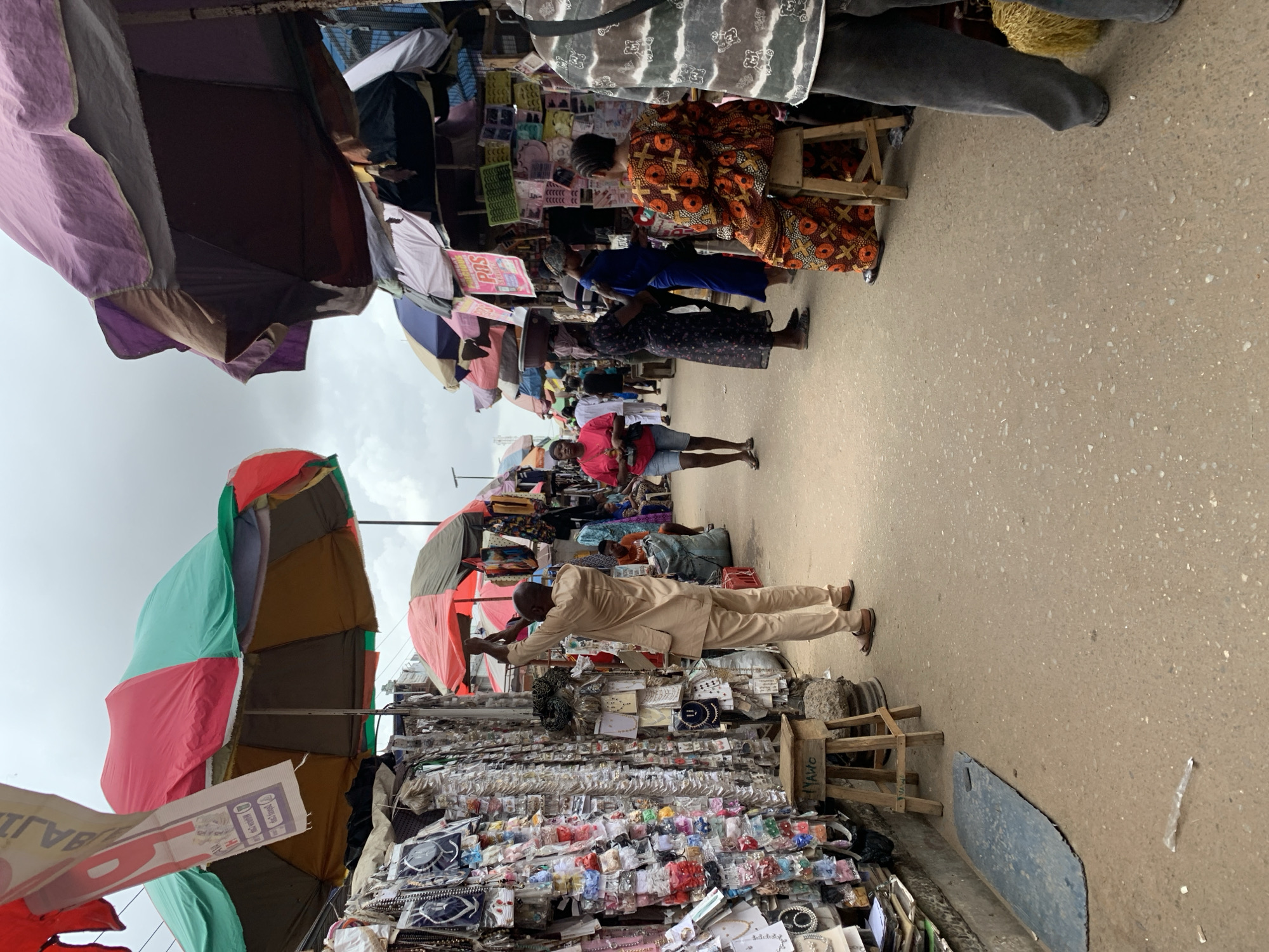The image shows a street scene in Ikorodu, Nigeria. A man in a light tan suit stands in front of a vendor stall displaying a large array of small items, possibly jewelry, hair accessories, and small trinkets. He is gesturing or holding something above the display. Overhead are numerous open umbrellas, providing shade for vendors and shoppers. Several people are walking through the market area. A woman is seated on a stool, while others are walking and wearing colorful clothes. The ground is paved.