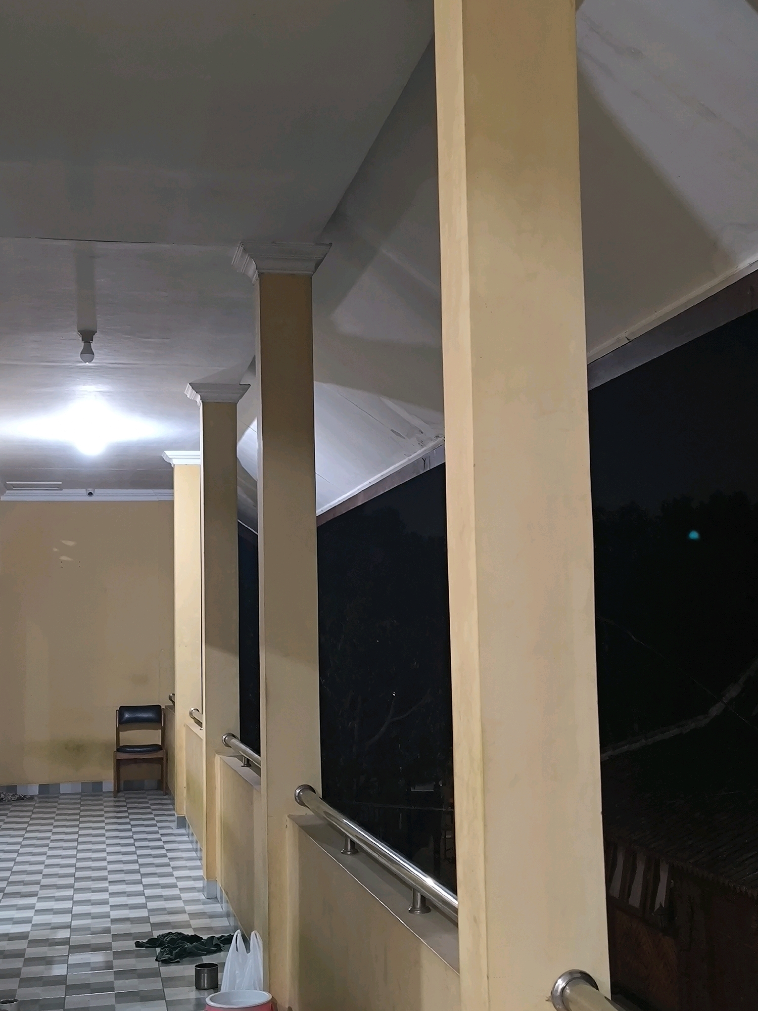 An exterior shot shows a covered walkway with yellow walls and a tiled floor. Several rectangular columns support the roof, which is illuminated by a bright light fixture. A black chair with a wooden frame sits against the wall on the left. On the floor, there are several items including a roll of dark fabric, a metal can, and a red and white bucket. A stainless steel railing runs along the edge of the walkway, separating it from the dark, tree-filled exterior.