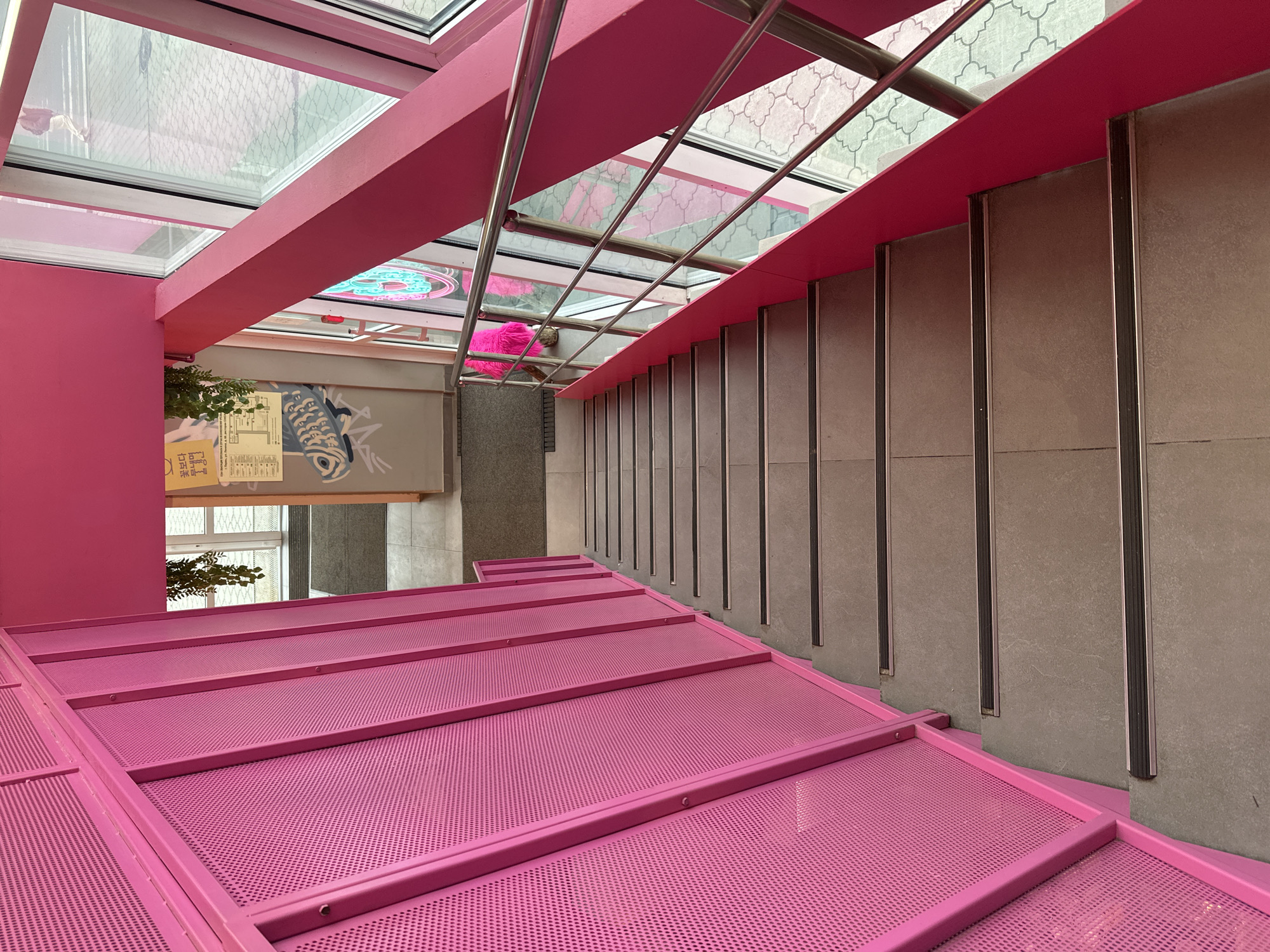 The image shows a downward-facing view of a bright pink staircase, likely indoors. The steps are made of a perforated metal material. To the right of the staircase are grey, vertical wall panels accented with black vertical strips. Above the staircase, there's a glass ceiling supported by a pink framework. A section of wall to the left is painted the same bright pink. Through the glass ceiling, a view of an exterior space is visible, and some interior elements like a neon sign and a pink, fur-like object can be seen. There is a gray wall behind the pink one with art work on it, including one of a fish. There is a green leafy plant sitting near the wall. The location is in the city of Perm, Russia.