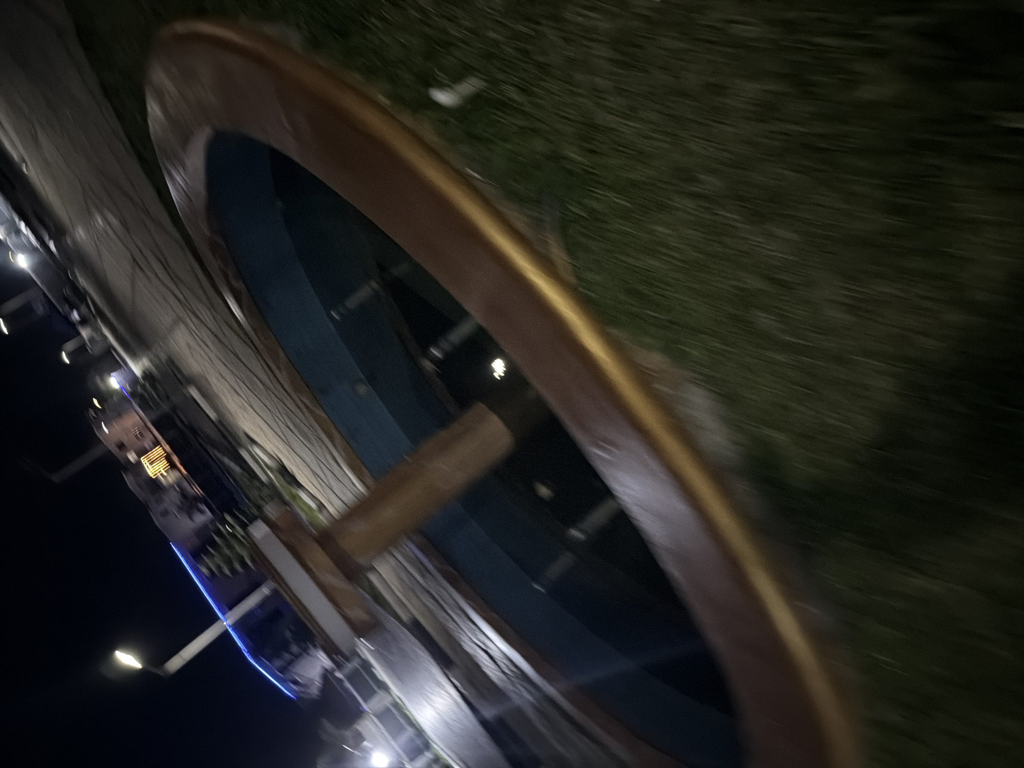 The image shows a night scene in Bukuru, Nigeria, featuring a fountain with a wooden rim and a blue interior, surrounded by grass. In the background, there are buildings with lights, streetlights, and blue lighting accents, suggesting a public or commercial area. The overall image is blurry due to motion.