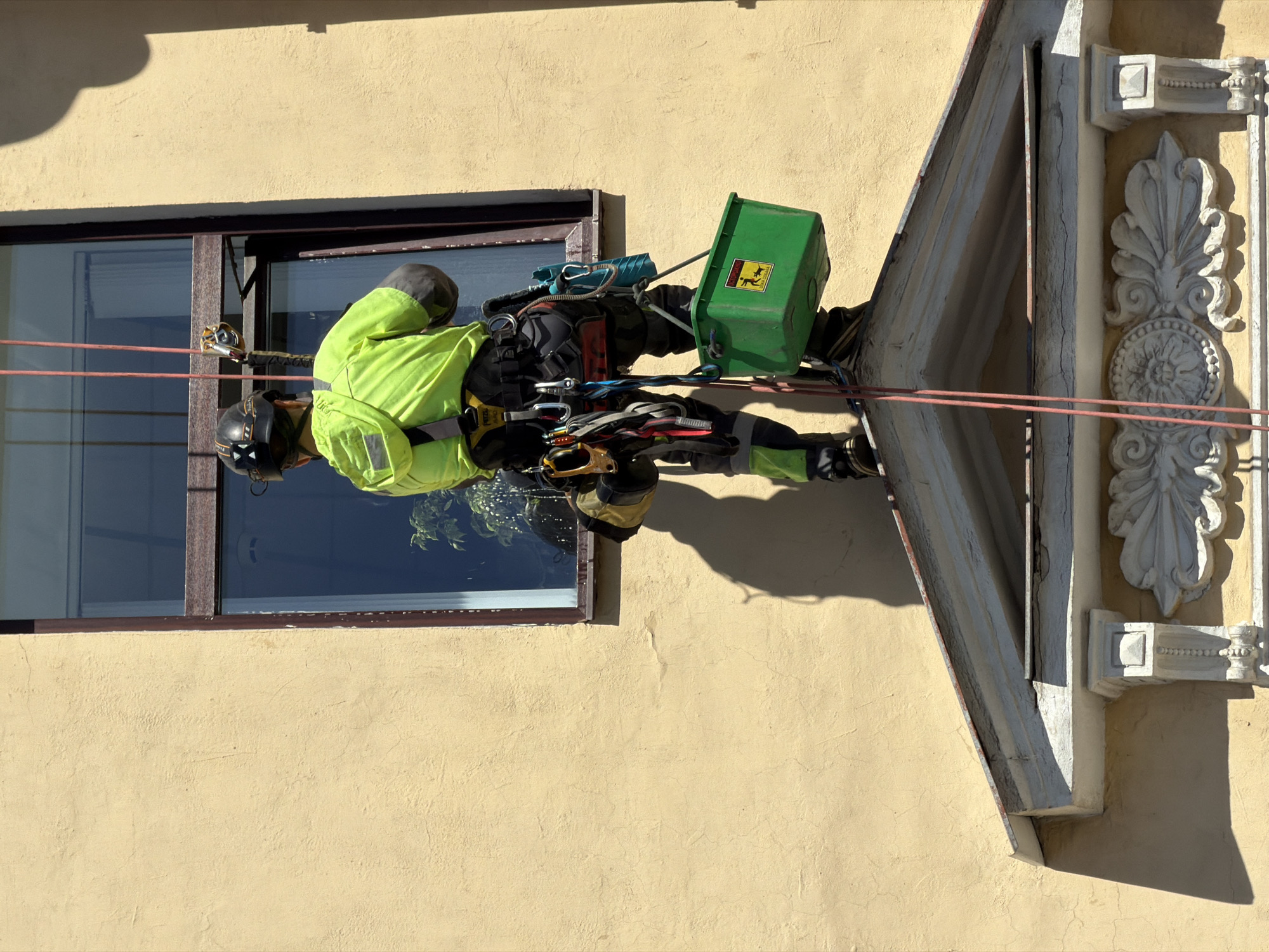 A lone individual is depicted from the back, suspended by ropes on the exterior of a light yellow stucco building under bright daylight. The person, appearing male, wears a neon green long-sleeved shirt with dark accents, dark trousers with neon green lower sections, and a black hard hat with an "X" symbol. A professional climbing harness, marked "PETZL," is worn, from which multiple carabiners and other rope access devices are attached. Two red ropes descend past the individual's left side. A green plastic bucket, hanging from the harness, features a yellow warning sticker showing a falling figure with the text "CAUTION FALLING OBJECTS." The individual's right hand holds a window cleaning squeegee, positioned against the glass of a dark-framed window.
Below the window, a white classical architectural pediment with ornate carvings, including volutes and a central medallion, projects from the facade. The individual is standing on the sloped roof of this pediment. The building's stucco facade shows minor cracks, indicative of age. Strong sunlight creates sharp shadows of the individual and the architectural features on the wall. Reflections in the window glass show parts of the sky and internal foliage. The architectural style, particularly the ornate classical pediment and stucco facade, is characteristic of historical buildings found in Saint Petersburg, Russia.