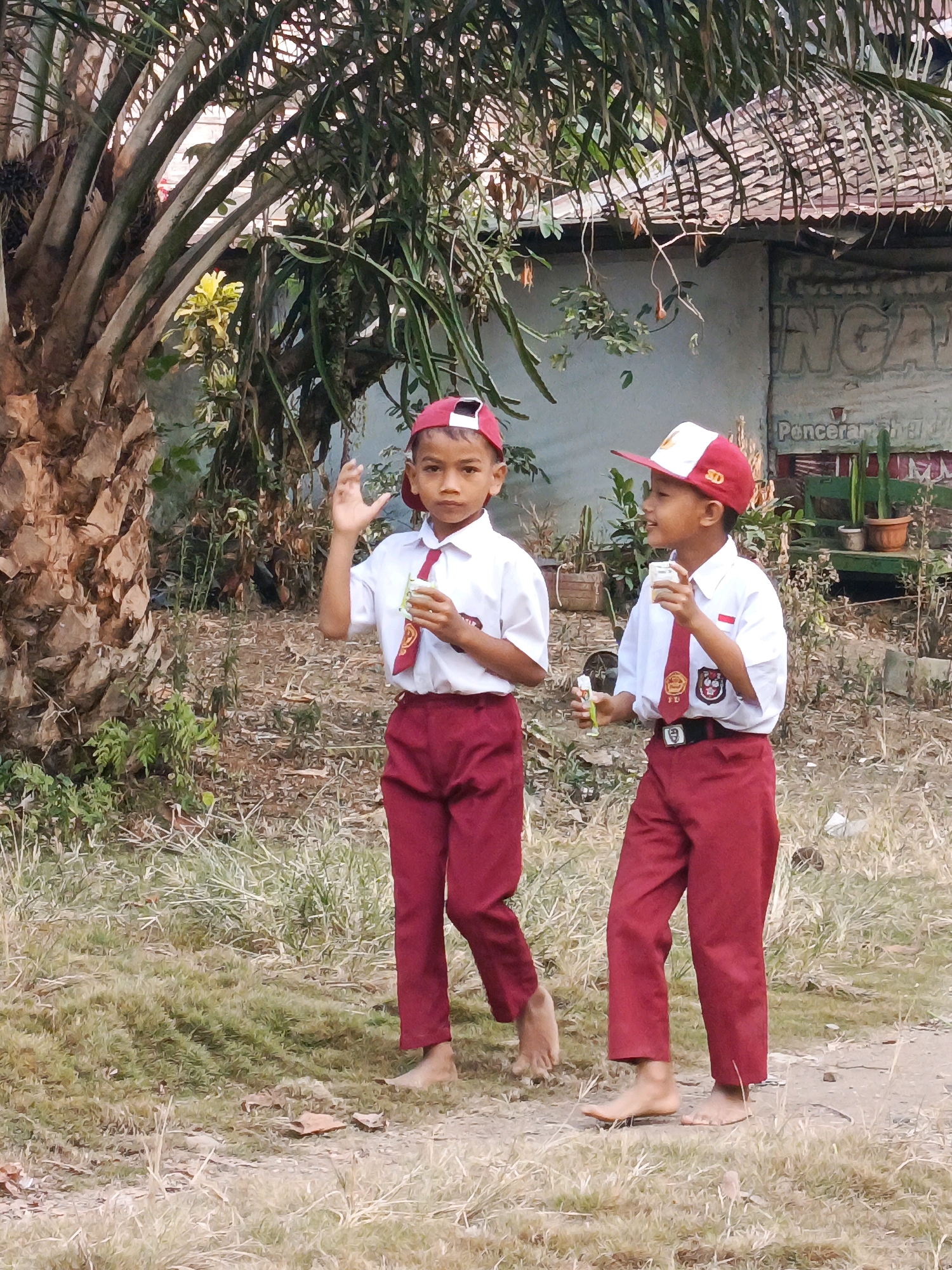 Two young boys, appearing to be elementary school students in uniform, stand in a grassy outdoor area with a large palm tree to their left. The boy on the left wears a red baseball cap, a white shirt with a red tie, and red pants. He is holding a small, clear plastic cup with a yellow lid in his right hand and waving his left hand. The boy on the right wears a red and white baseball cap, a similar white shirt with a red tie, and red pants. He is holding a small, clear plastic bottle with a green cap in his left hand. Behind them, a building with a corrugated metal roof is visible, with a sign that reads "NGA" and "Poncera..." further to the right. There are potted plants and dry vegetation surrounding the area. The ground is covered in dry grass and some dirt. The scene suggests a casual setting, possibly near a school or residential area in Megang Sakti II, Indonesia.