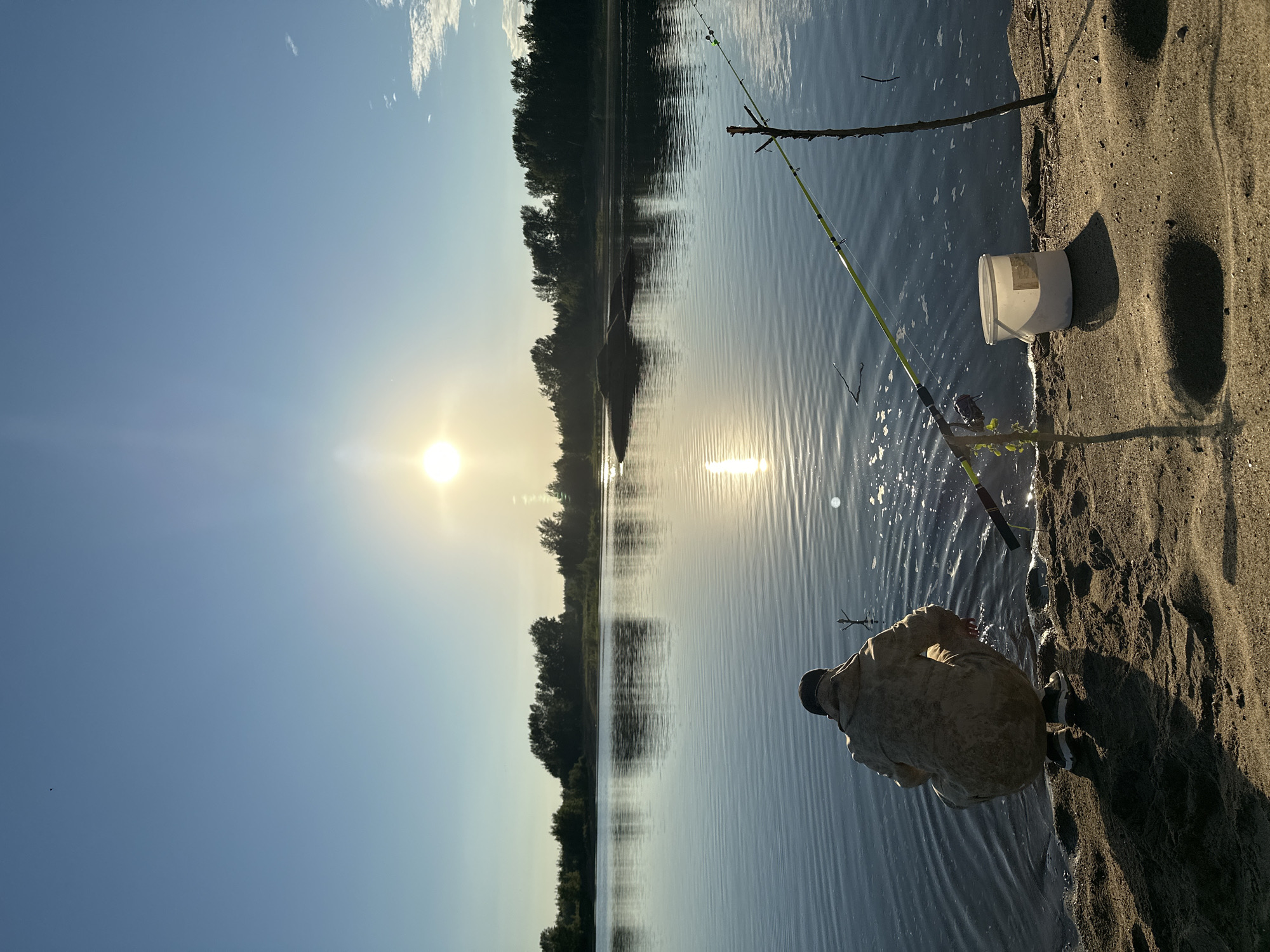 A person is crouched on a sandy bank, facing a calm body of water. The sun is low in the sky, casting a bright glare on the water's surface and creating reflections. A fishing rod is propped up on the bank, its line extending into the water. A white bucket sits near the fishing rod. Trees line the opposite bank of the water, forming a dark silhouette against the bright sky. The location is the city of Noviy, Russia.