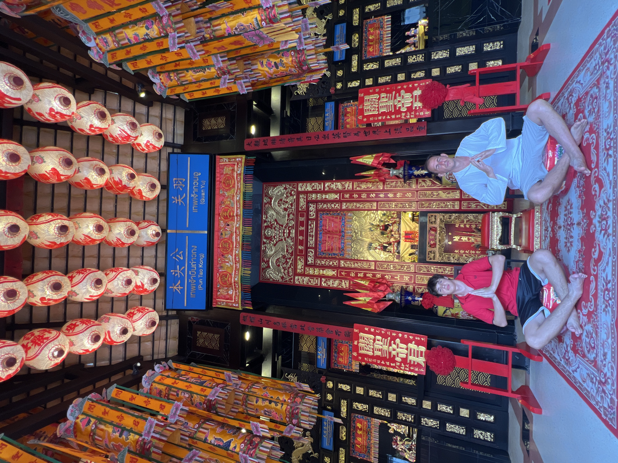 Two men are meditating in front of a shrine at the Pun Tao Kong Temple in Baan Hua Thanon, Thailand. The man on the left wears a red polo shirt and black shorts, while the man on the right wears a white long-sleeved shirt and white shorts. Both men are seated cross-legged on red cushions placed on a patterned red rug, with their hands pressed together in front of their chests. The shrine features ornate red and gold decorations, including a large red altar with golden dragon motifs, and intricate wooden panels on the walls. The ceiling is adorned with numerous paper lanterns in red and white, and vibrant yellow banners with Chinese calligraphy hang down the sides of the shrine. The overall environment is one of cultural and spiritual significance, with a focus on traditional Thai and Chinese aesthetics.