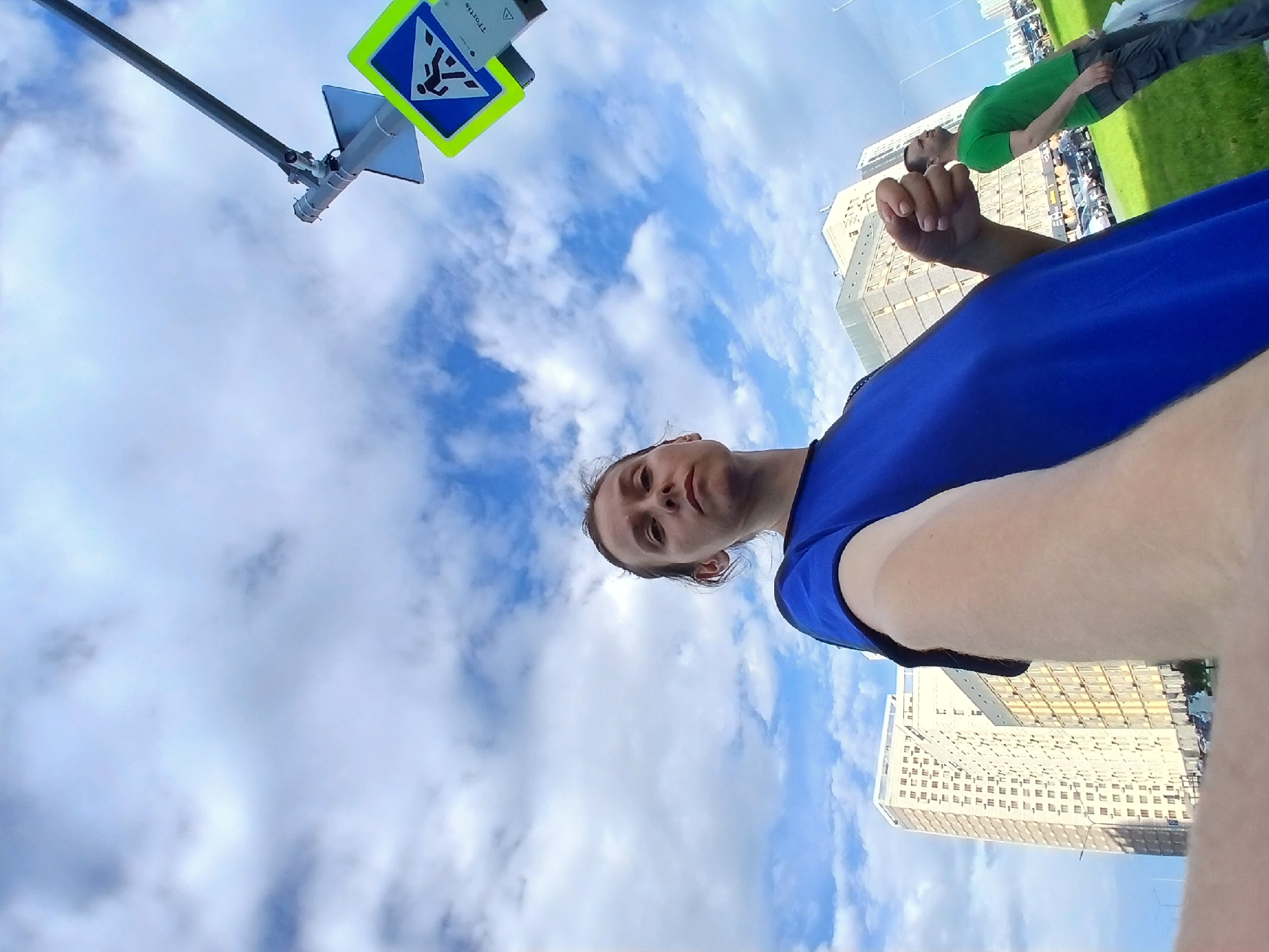 A first-person perspective selfie shows a woman in a blue shirt, her left arm extending towards the camera. Behind her, to the right, a man in a green shirt and grey pants stands looking up. In the background, tall apartment buildings are visible under a cloudy sky. A pedestrian crossing sign is mounted on a pole to the right of the woman. The scene appears to be in a modern urban area, possibly Yekaterinburg, Russia, based on the architecture and signage.