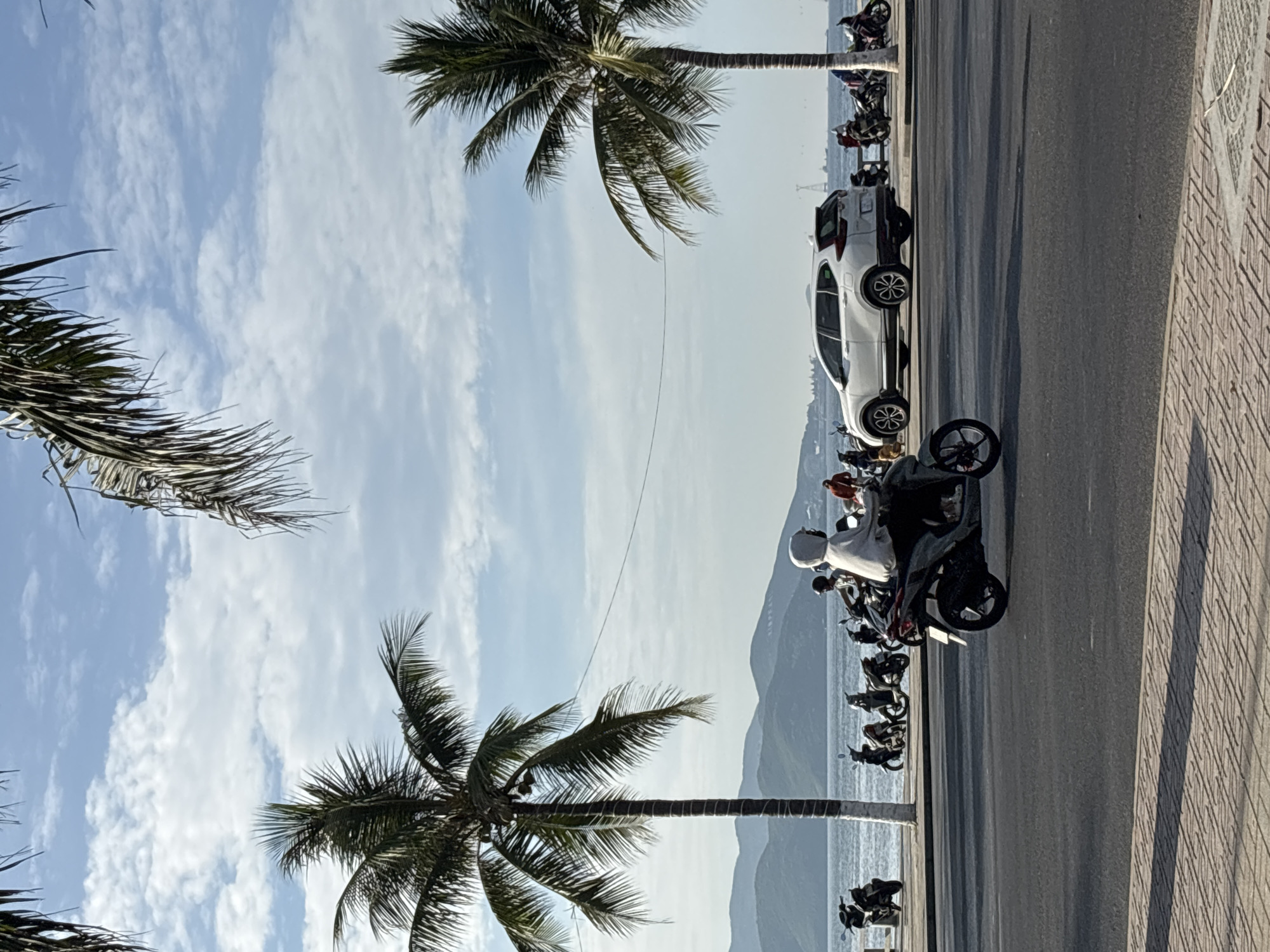 This is a street-level, eye-level, medium-shot photograph taken in Nha Trang, Vietnam, likely along a coastal road near the Tran Phu Bridge area. The foreground shows a paved road with a patterned brick sidewalk. In the middle ground, a motorbike with a rider wearing a white hoodie and helmet is traveling to the left. To the left of the motorbike, a line of parked motorbikes is visible along a low wall. Behind the parked motorbikes is the ocean, with a distant view of lush green mountains under a partly cloudy sky. Two prominent palm trees frame the scene, one on the left and another on the right. A white SUV is parked to the right of the line of motorbikes, facing right. The sky is a clear blue with scattered white clouds. The overall scene depicts a typical day in a Vietnamese coastal city.