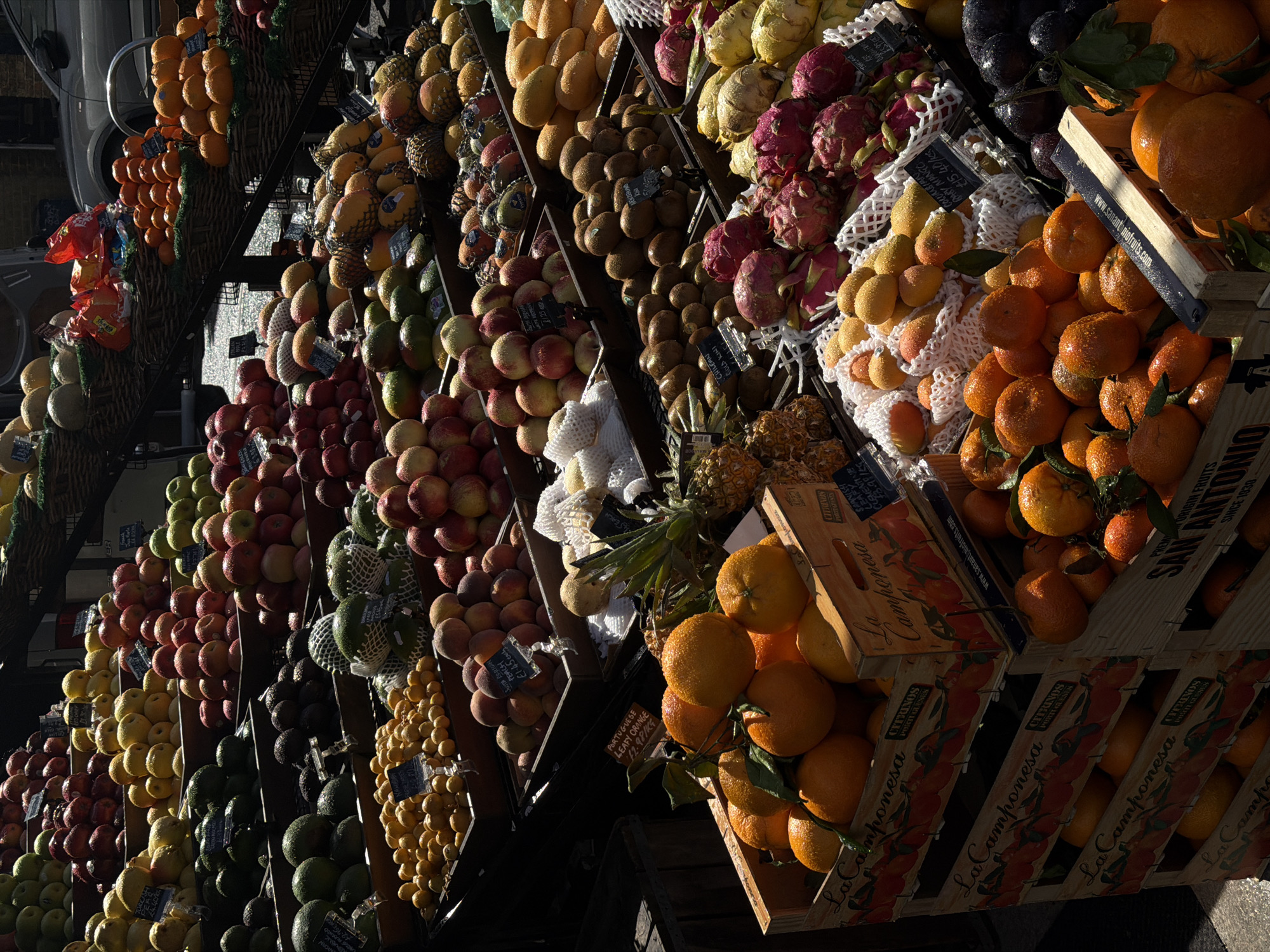 The image depicts a vibrant fruit stand in what appears to be an outdoor market, located possibly in London, United Kingdom, judging by the text on the price tags and crates. Several types of fruits are neatly arranged in baskets and wooden crates, cascading down a multi-tiered display. Oranges, apples, peaches, avocados, kiwis, dragon fruit, mangos, and other fruits are visible. Price tags are attached to some of the fruit displays. The wooden crates have text in Portuguese and English, indicating the origin of the fruit and the name of the distributor or producer. The angle of the image suggests a low viewpoint, looking upwards at the stacked fruit displays. Shadowy areas are prominent in the lower part of the image.