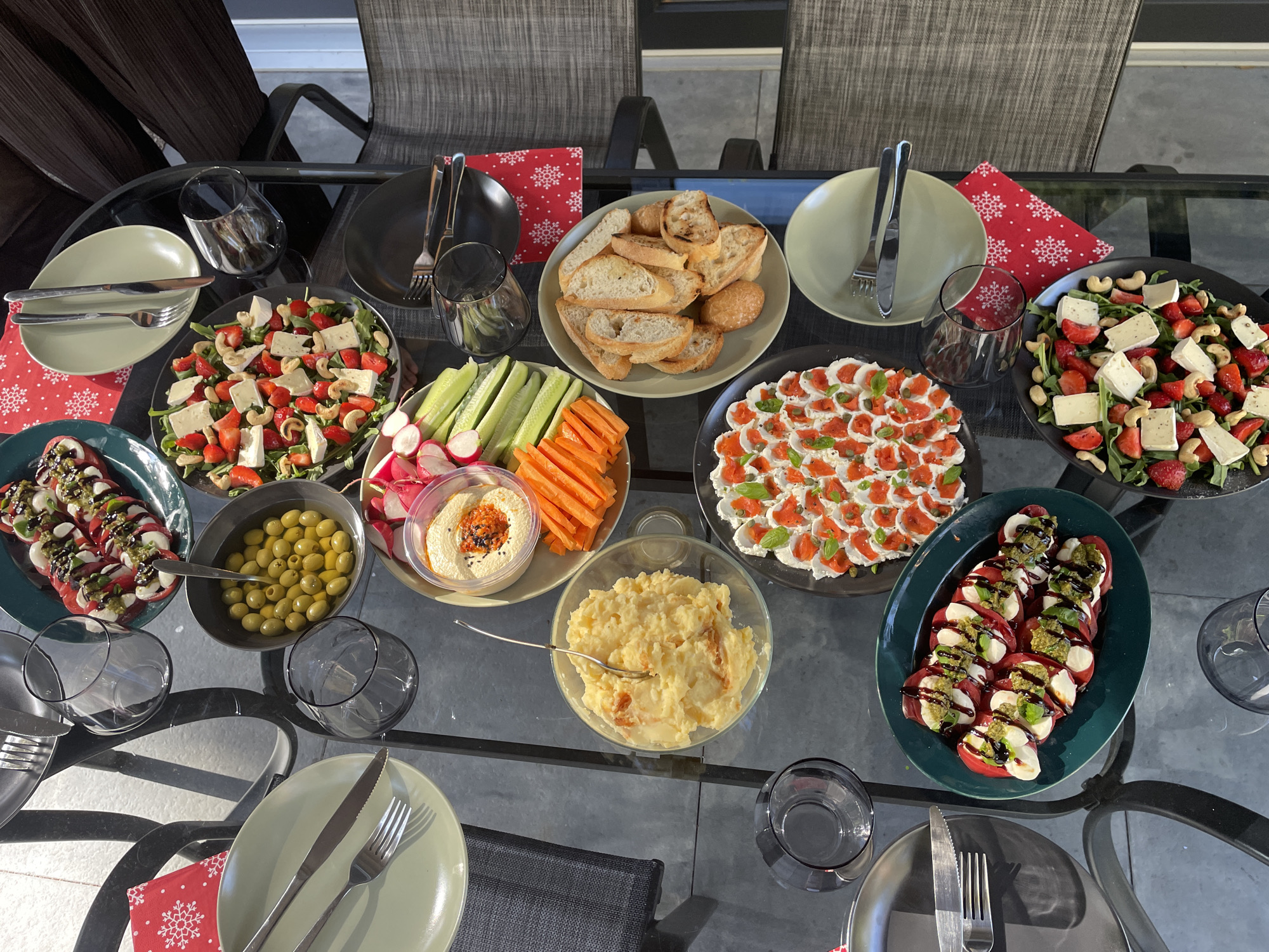 A top-down, high-angle medium shot shows a glass-topped outdoor dining table laden with various appetizers and drinks. The table is set with plates, silverware, and glassware, suggesting a meal in progress. The appetizers include a Caprese salad with sliced tomatoes, mozzarella, and basil, a bowl of green olives, a platter of sliced bread, a bowl of mashed potatoes with some visible pieces of what appears to be roasted red pepper or paprika mixed in, a serving of hummus with crudités (carrots, cucumbers, and radishes), and a strawberry and brie salad with cashews and arugula. Several glasses, some containing red liquid, are placed around the table. Red napkins with white snowflake patterns are tucked under some of the place settings. The scene is set outdoors, as evidenced by the sunlight casting shadows and the presence of patio furniture.