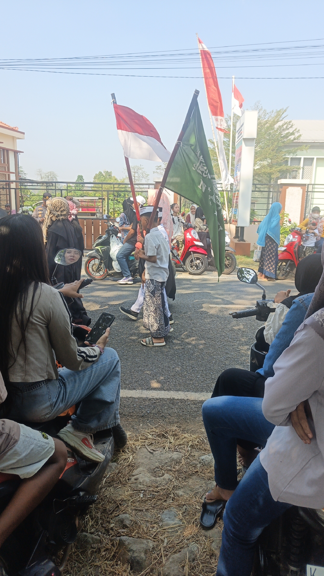 In a street scene under a clear blue sky, a group of people are gathered, many on or around motorcycles. Two flags are prominently displayed: one is the Indonesian flag, red and white, and the other is a green flag with Arabic script.
In the foreground, a woman with long dark hair is seated on a motorcycle, looking at her phone. To her left, another person's legs are visible, wearing light-colored shorts and a patterned fabric.
In the mid-ground, a young boy in a white cap and patterned pants holds one of the flags. Other individuals are visible standing and seated, some wearing traditional Islamic attire like hijabs and abayas. Several motorcycles are parked or being ridden, including a bright red scooter.
The background shows buildings with light-colored facades and tiled roofs, along with trees and a fence. The ground is asphalt, with some dry vegetation and stones scattered near the edge of the road. The overall impression is of a community gathering or event taking place outdoors.