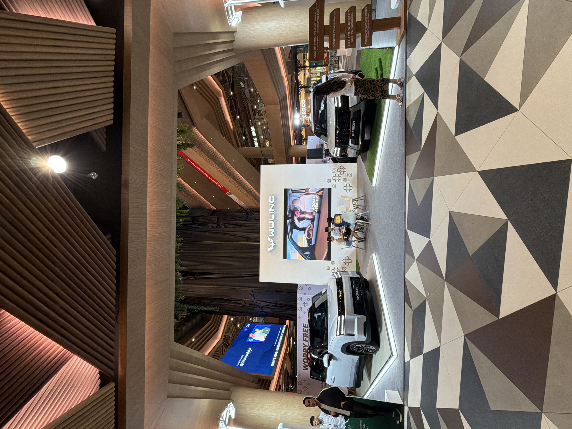 This image captures an indoor car exhibition or showroom, likely within a modern shopping mall or convention center in Denpasar, Indonesia. The scene is well-lit, suggesting daytime or evening hours under artificial light.
In the foreground, a white Wuling car model, specifically an "EKSION," is displayed on a raised platform with glowing perimeter lights. Another dark-colored Wuling "EKSION" car, with "CROCO" visible on its front, is also on display further back. The cars are presented in a promotional setting with signage highlighting features such as "ROOMY 4-SEATER," "7-SEATER CONFIGURATION," "1208- KM TOTAL COMBINED RANGE (CLTC)," "LIFETIME WARRANTY (Core PHEV Components)," and "MUSCULAR FLOWLINE DESIGN." A slogan "WORRY FREE" is also visible.
Several people are present, including a woman standing near the dark-colored car, another woman interacting with a display, and a few individuals seated at a small table near a screen displaying car interiors. Two men are visible in the lower foreground, one appearing to be a staff member.
Large digital screens are prominent. One screen associated with the Wuling display shows an interior view of a car, along with the brand "WULING" and various decorative patterns. Another large screen in the background displays text including "Kanal Youtube @Ditjenpajakri" and "TODAY'S NEWS: PAMERAN LUKISAN AT GROUND HAL," indicating other events or information being promoted in the venue. The name "RUNG MINA" is visible on a sign in the background.
The architecture features light-colored wood paneling, including striking slatted designs on walls and ceilings, creating a contemporary and inviting atmosphere. The flooring in the foreground has a distinctive geometric pattern of triangles in shades of grey, brown, and cream. Artificial greenery is used as part of the decor.