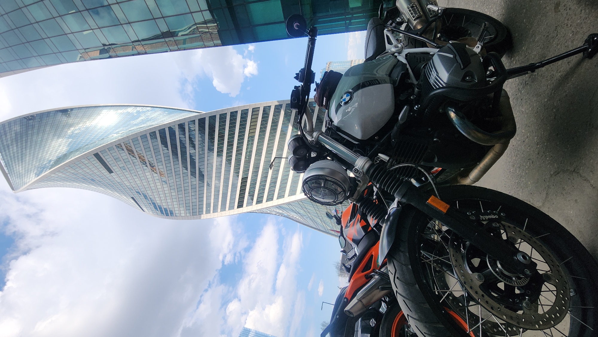 The image shows two motorcycles parked in front of a twisting skyscraper under a partially cloudy sky. The motorcycle in the foreground is primarily black and orange. Adjacent to it is a BMW motorcycle, notable for its gray and white color scheme and distinctive round headlight. The skyscraper in the backdrop is the Evolution Tower, situated in the Moscow International Business Center (Moscow-City) in Moscow, Russia.