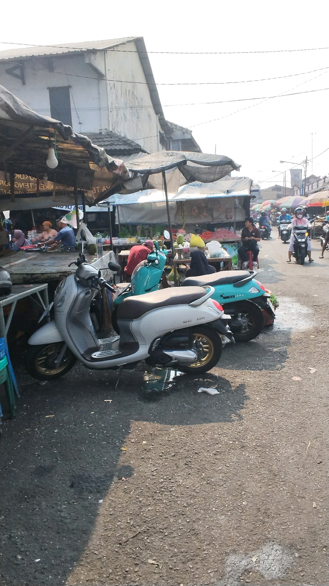 This image depicts a bustling market scene in Mojokerto, Indonesia, likely a roadside market given the presence of parked motorcycles and a street surface. In the foreground, two scooters are prominently displayed: a silver scooter on the left and a teal scooter to its right. Behind the scooters, market stalls are set up under tarpaulin canopies, displaying various goods, possibly produce, judging by the colorful items visible. Several individuals are present, some tending to the stalls, while others are seen walking or riding motorcycles in the background. The architecture visible in the background suggests a mix of simple buildings, characteristic of a local market area. The lighting indicates it's daytime, with sunlight casting shadows on the ground.