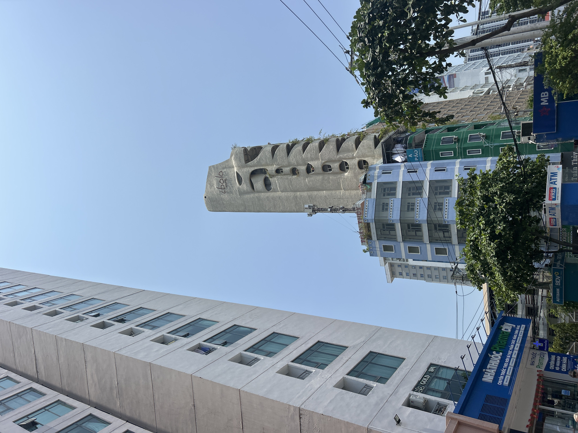 The image captures a street scene in Nha Trang, Vietnam, featuring a mix of modern architecture and local businesses. On the left, a white building with horizontally arranged rectangular windows is visible. Above it, the unique architectural design of the "ibala Hotel" stands out, characterized by its concrete structure with rounded openings and green foliage on top. In the background, a blue-and-white building and a green building can be seen. Street signs and advertisements for local businesses, including a pharmacy ("Pharmacity") and a bank ("BIDV"), line the side of the road. Utility wires crisscross the sky, and trees provide some greenery to the urban landscape.