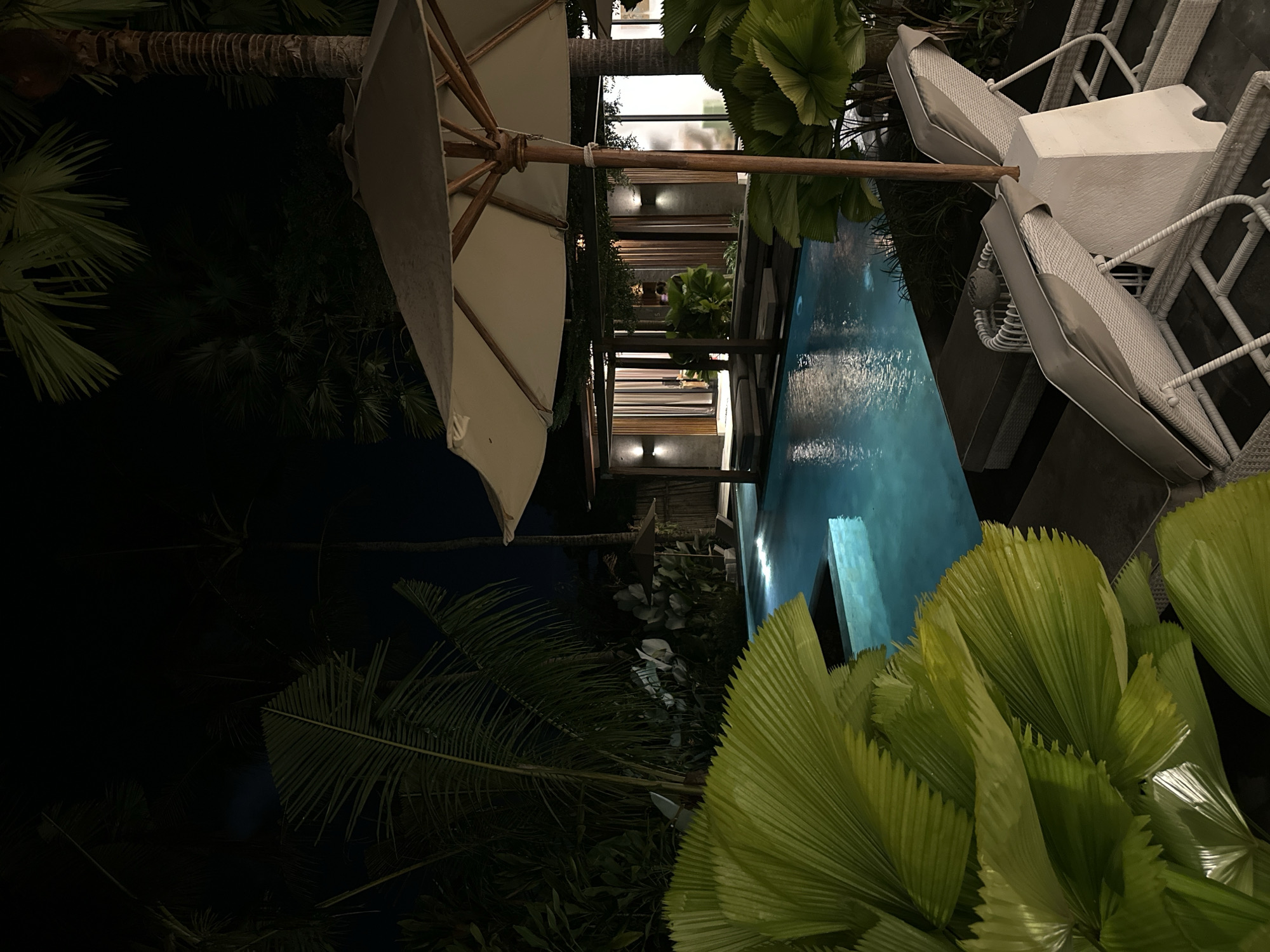 The image depicts a nighttime scene at a location in Sukawati, Indonesia, potentially a resort or private residence featuring a swimming pool. A partially opened white patio umbrella dominates the upper left portion of the frame. The pool's blue water reflects light, suggesting underwater or surrounding illumination. Adjacent to the pool are white patio chairs with grey cushions and a small square white table. Lush tropical vegetation, including palm trees and fan-shaped plants, surrounds the pool area, contributing to the tropical ambiance. The background shows a building with multiple levels, illuminated by warm lighting, possibly indicating an interior or walkway. The overall atmosphere suggests a luxurious and tranquil setting.