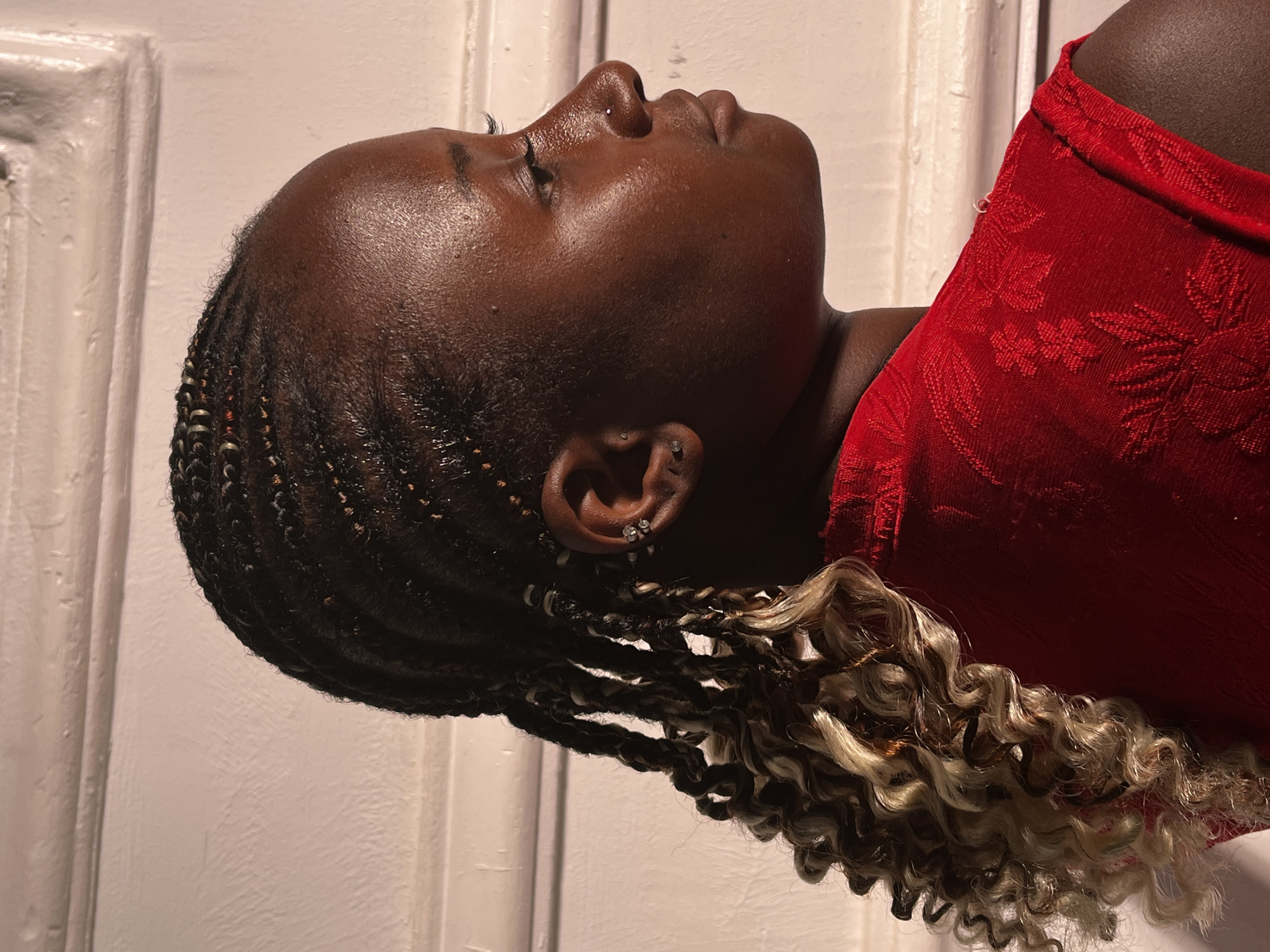 A person with dark skin is facing upwards and to the right of the image wearing a red garment with floral embroidery near a white wall. The individual's hair is braided with blonde extensions near the ends. They have earrings in their left ear. The location is unknown, and there is no indication it is in Barakin Akawo, Nigeria.