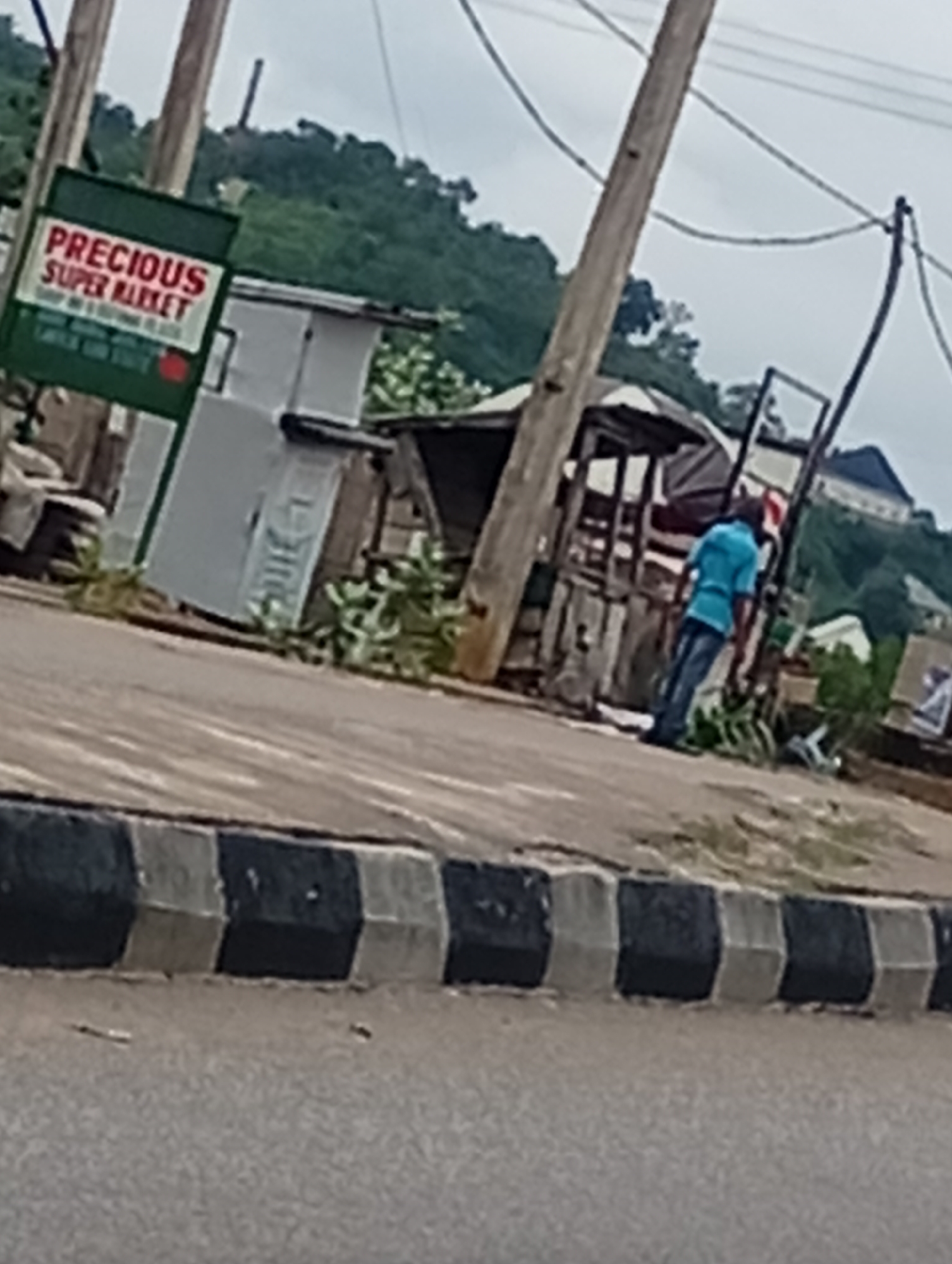 A person in a blue shirt and jeans stands on a dirt road in Adankolo, Nigeria. They are facing away from the camera, with a small, dark-colored structure behind them and a green sign reading "PRECIOUS SUPER MARKET" to their left. The sky is overcast, and the ground is a mixture of dirt and pavement. In the background, lush green hills rise, with a white building visible on the hillside. The scene appears to be in a semi-rural or suburban area.