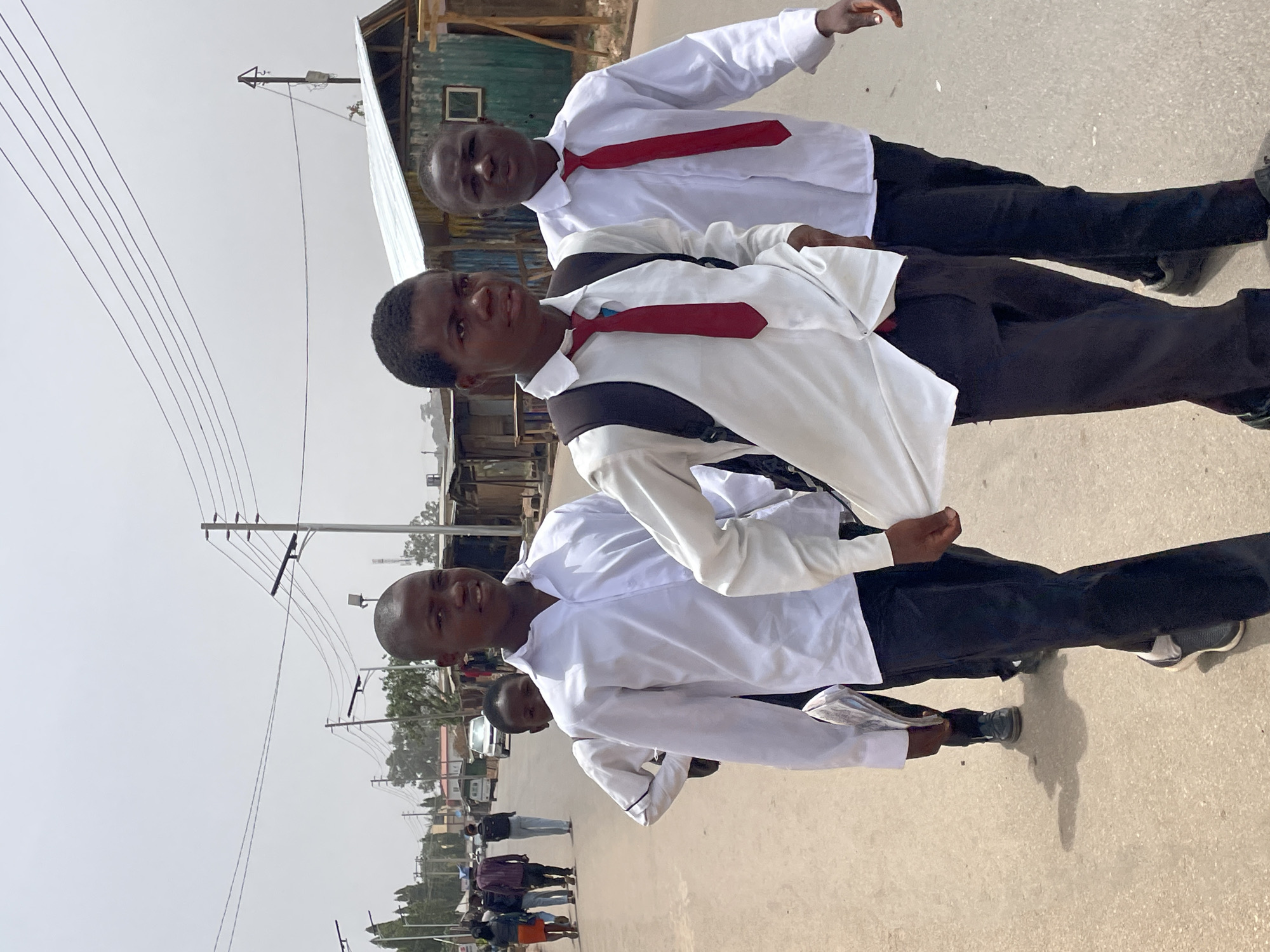 The image shows three young men walking in what appears to be Jos, Nigeria, based on the architecture and infrastructure visible. They are wearing white shirts, black pants, and red ties, suggesting school uniforms. The environment is an urban street with overhead power lines and buildings in the background. Other people walk along the road in the background. The men are facing the camera.