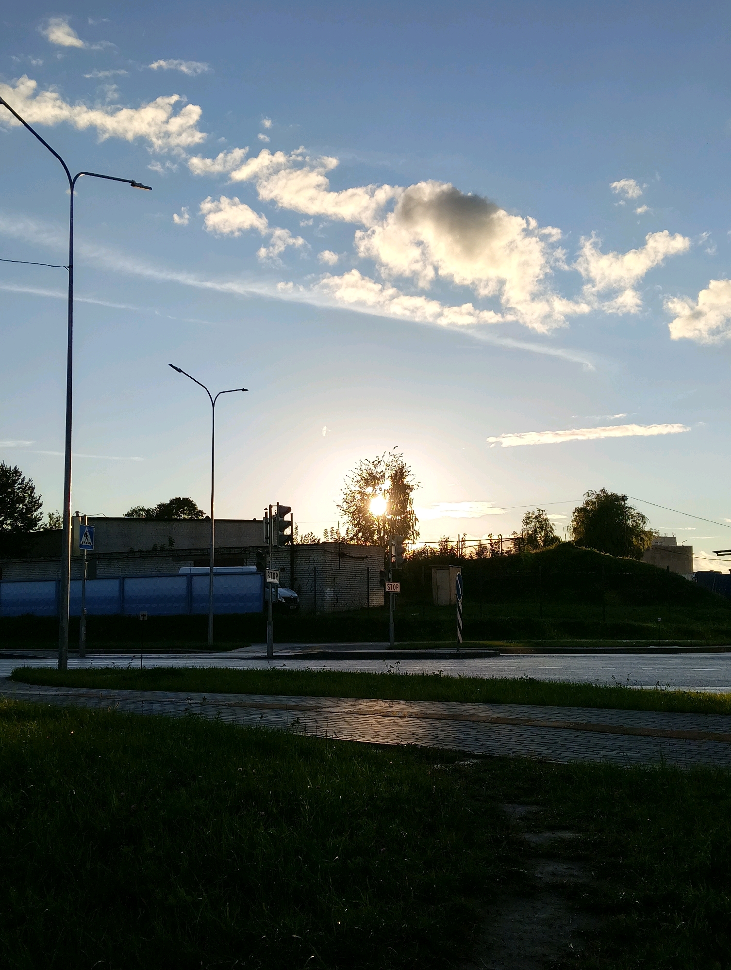 This image captures an outdoor scene with a clear sky and scattered clouds, illuminated by the sun which is low on the horizon, suggesting it is either sunrise or sunset. The foreground features a patch of dark green grass and a paved walkway. In the mid-ground, there is a road with a slight sheen, indicating recent rain or dampness, and a grass verge. Beyond the road, a light-colored brick building and a darker, lower fence line are visible. Several streetlights are present throughout the scene. A traffic signal with a "STOP" sign is mounted on a pole. Trees with green foliage are scattered across the landscape, with some appearing to be silhouetted against the bright sky. The environment appears to be an urban or suburban setting.