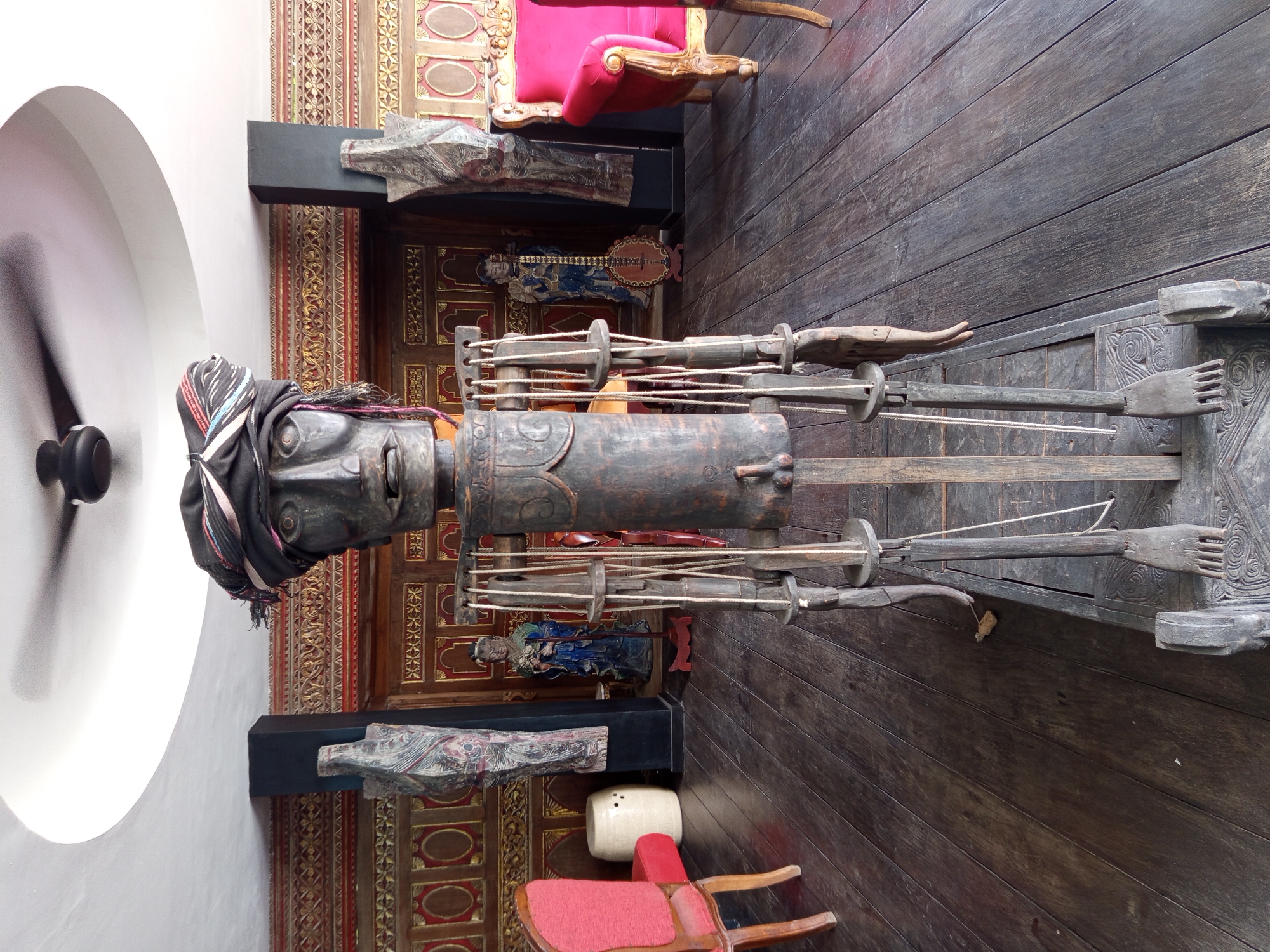 The image shows a wooden puppet figure, likely part of a traditional performance art, positioned in front of an ornate wall with gold detailing. The figure is mostly dark wood with a carved face and a cloth headdress. It has long wooden arms and hands attached to a supporting structure with strings. The base is decorated with carvings. The floor is made of dark wood planks. Several pieces of furniture are also present, including red chairs and a carved wooden figure on the wall. The image was taken in North Kuta, Indonesia.