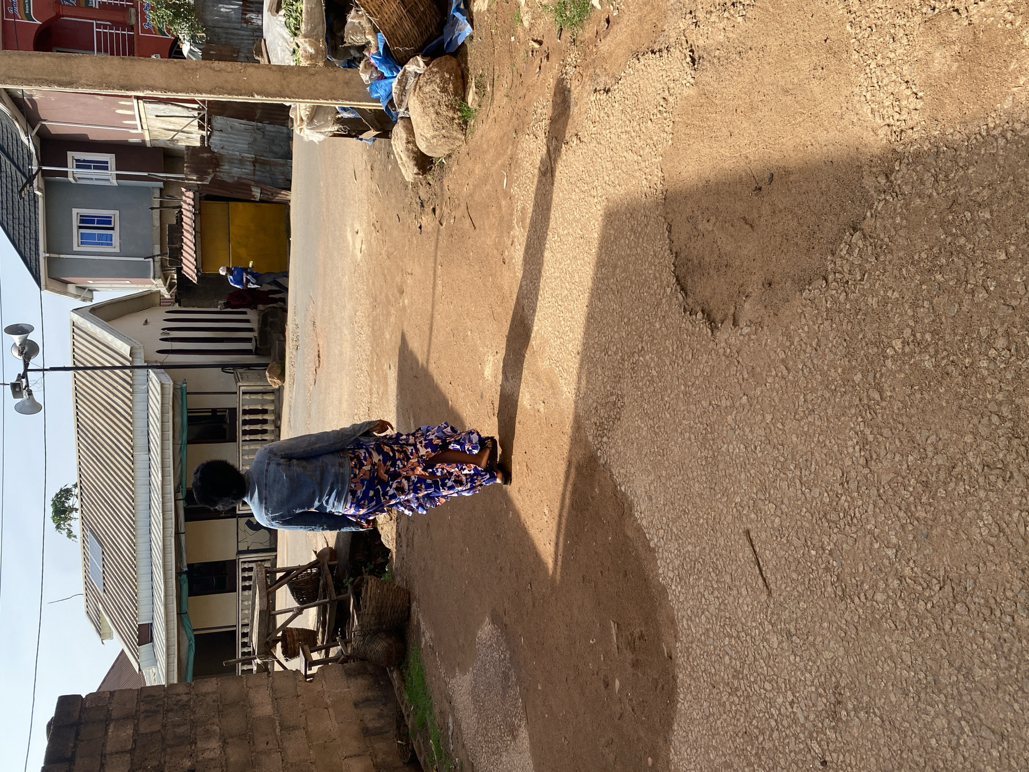 A rear view of a woman with dark, curly hair wearing a denim jacket and a long, ruffled floral skirt walks away from the camera on a dirt and gravel road. She is in the foreground, casting a long shadow. To her left, a brick wall and a wooden structure with baskets are visible. In the background, several buildings are present, including one with a light-colored facade and a corrugated metal roof, and another with a gray and maroon exterior. Two individuals in the mid-ground are walking on the road. The scene appears to be a street in Jos, Nigeria, characterized by its unpaved road, building styles, and a generally dusty environment. The sky is clear and blue, suggesting a sunny day.
