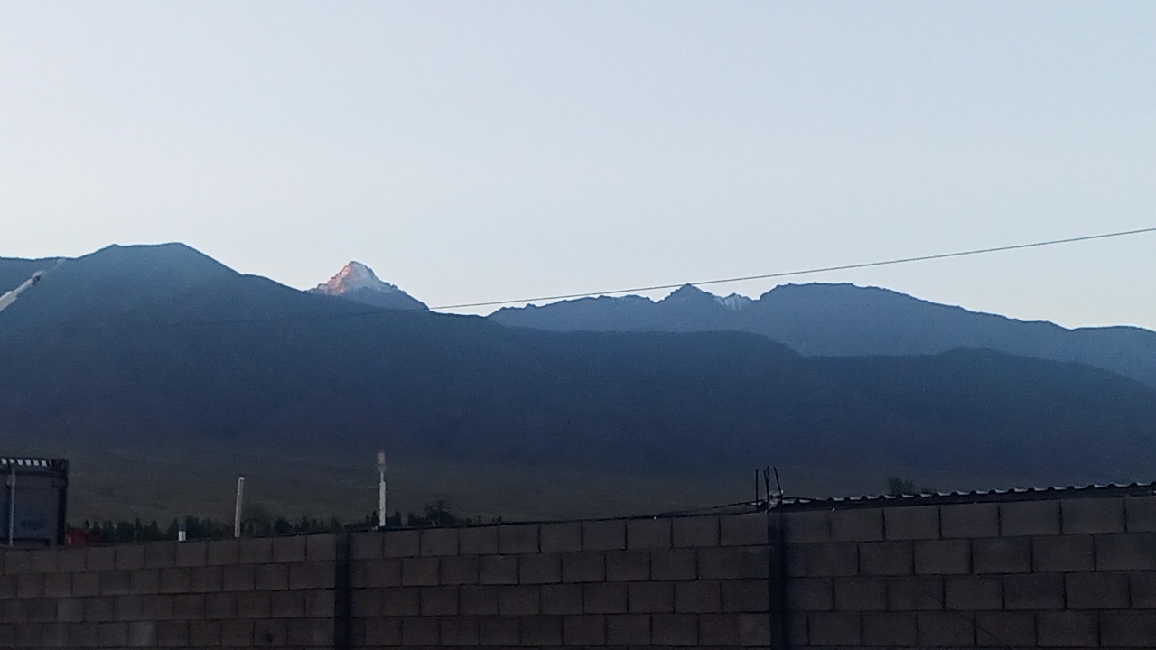 The image depicts a panoramic view of dark, silhouetted mountain ranges against a pale sky, likely at dawn or dusk. The mountains exhibit layered contours, with some peaks appearing snow-capped. In the foreground, a brick wall runs horizontally across the bottom of the frame, partially obscuring a landscape of sparse vegetation and utility poles. The lighting suggests an outdoor scene in a natural setting with man-made structures in the foreground. The location is identified as Choktal, Kyrgyzstan.