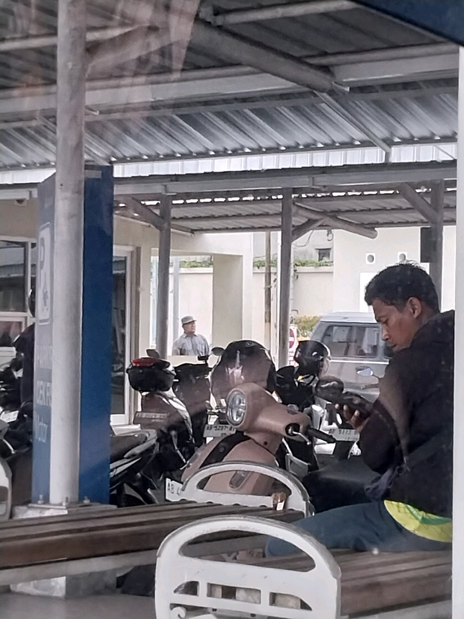 A man wearing a dark jacket and blue pants is sitting on a wooden bench, looking at his phone. He is positioned in the foreground on the right side of the image, with his body angled to the left. In the background, several motorcycles are parked under a covered structure with a corrugated metal roof. Another person wearing a light-colored cap and shirt stands in the middle ground, looking to the left. A silver van is visible behind the motorcycles. The scene appears to be an outdoor parking area, possibly at a public facility in Sleman, Indonesia, given the presence of motorcycles and a van. The lighting suggests it is daytime.