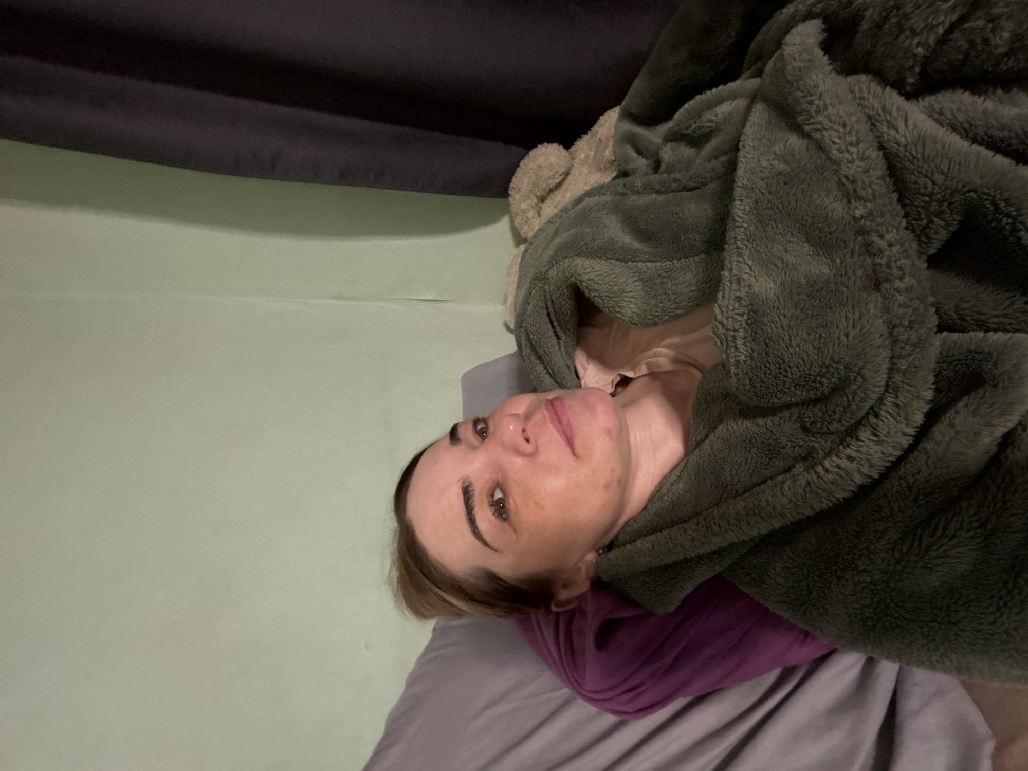 A woman with light skin and brown hair is lying down on a bed with gray sheets and a purple pillow. She is partially covered by a green, fuzzy blanket. The background consists of a light green wall and a dark gray curtain or drapery. The location is unknown.