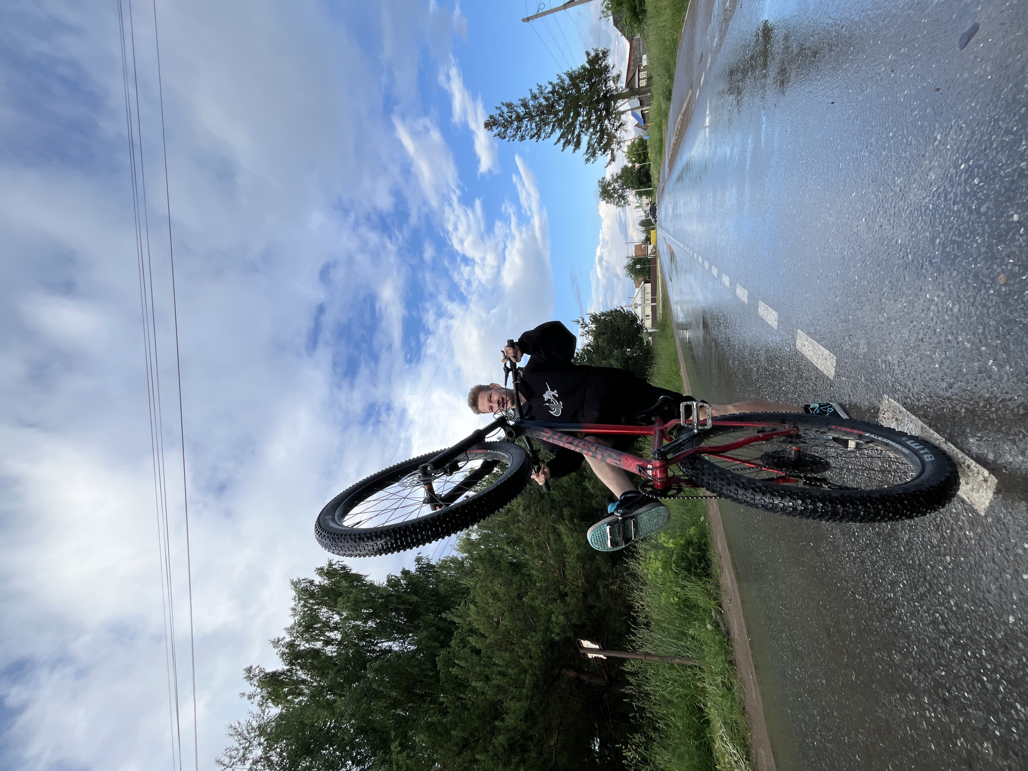 A young Caucasian male with blond hair is performing a wheelie on a red bicycle on a wet asphalt road. He is wearing a black hoodie with a white graphic and black pants. The bicycle has black tires and a red frame. The road has a white dashed line and is reflecting the sky, indicating it is wet. The scene is in a rural or suburban area with trees and houses on either side of the road. The sky is cloudy with patches of blue. The image appears to be taken in the city of Chaikovsky, Russia, likely during daytime.