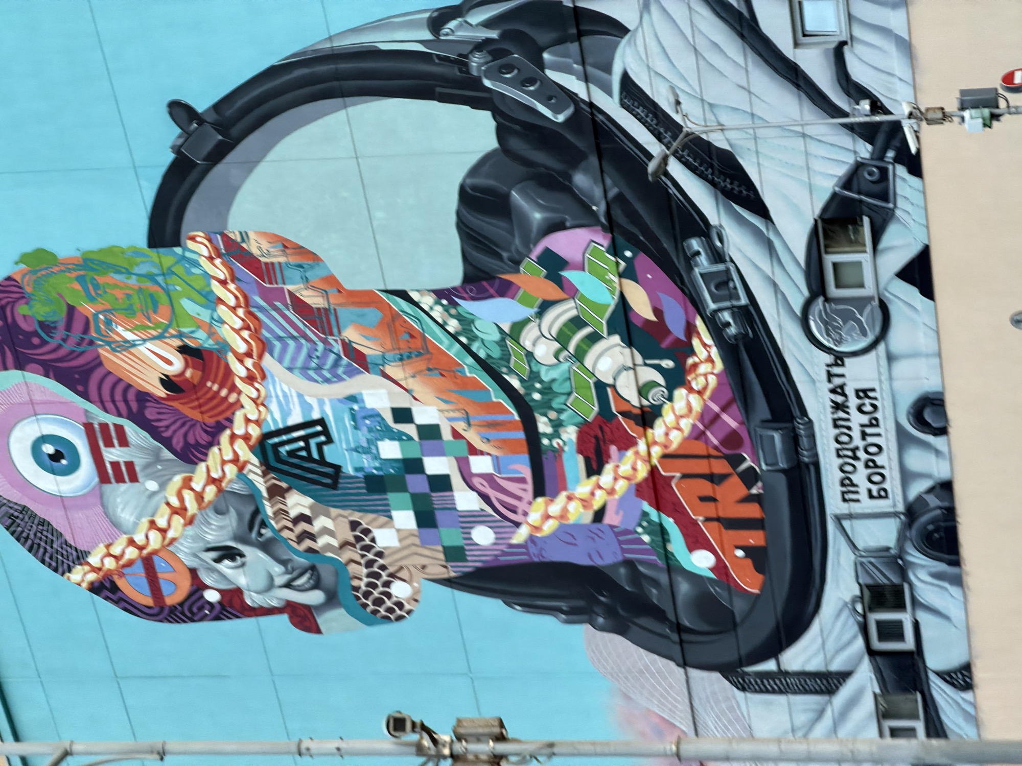The image shows a mural on a building in Moscow, Russia. The mural depicts a stylized astronaut helmet with vibrant, colorful patterns and designs within the helmet's visor. The helmet is predominantly black and white, with the interior of the visor filled with a collage of images, including a woman's face, an eye, and various geometric shapes. A gold chain is draped across the visor. The helmet is attached to a spacesuit that has "ПРОДОЛЖАТЬ БОРОТЬСЯ" (CONTINUE TO FIGHT) written on a strap. The background is a light blue. The mural is located on the side of a building, as evidenced by the presence of windows and a utility pipe running in the foreground.
