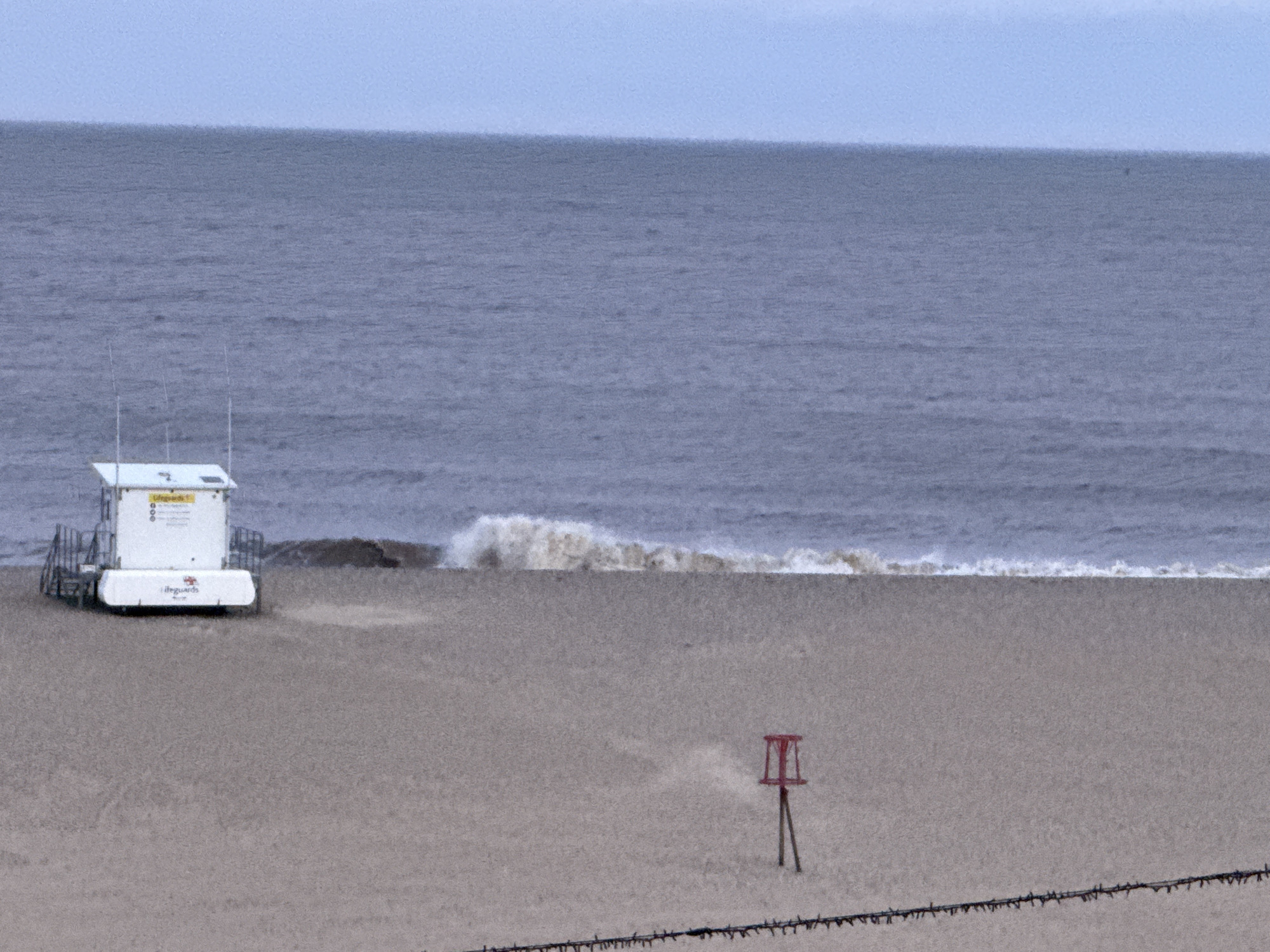 The image depicts a beach scene in Gorleston-on-Sea, United Kingdom. A white lifeguard station stands on the sand near the water's edge. A red channel marker is visible further down the beach. Waves are breaking in the background. The beach appears mostly empty. A wire fence runs along the bottom of the image. The sky is overcast.