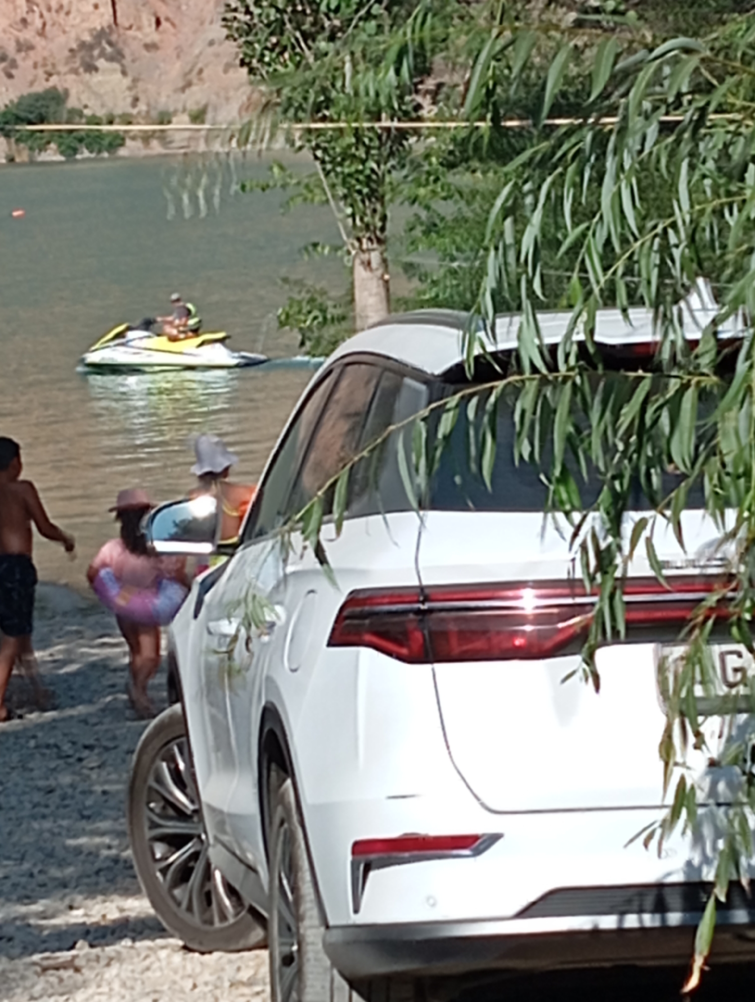 A white SUV is parked on a rocky shore next to a body of water. In the water, a person is riding a jet ski. On the shore, a child in black shorts stands with their back to the camera, facing the water. To the right of the child, a young person wearing a pink hat and a pink inflatable ring walks towards the water. To the right of them, another person wearing a white hat and a yellow top is visible. The environment includes a rocky cliff face in the background, trees, and foliage obscuring parts of the SUV. The scene is set in Yangikurgan, Uzbekistan.