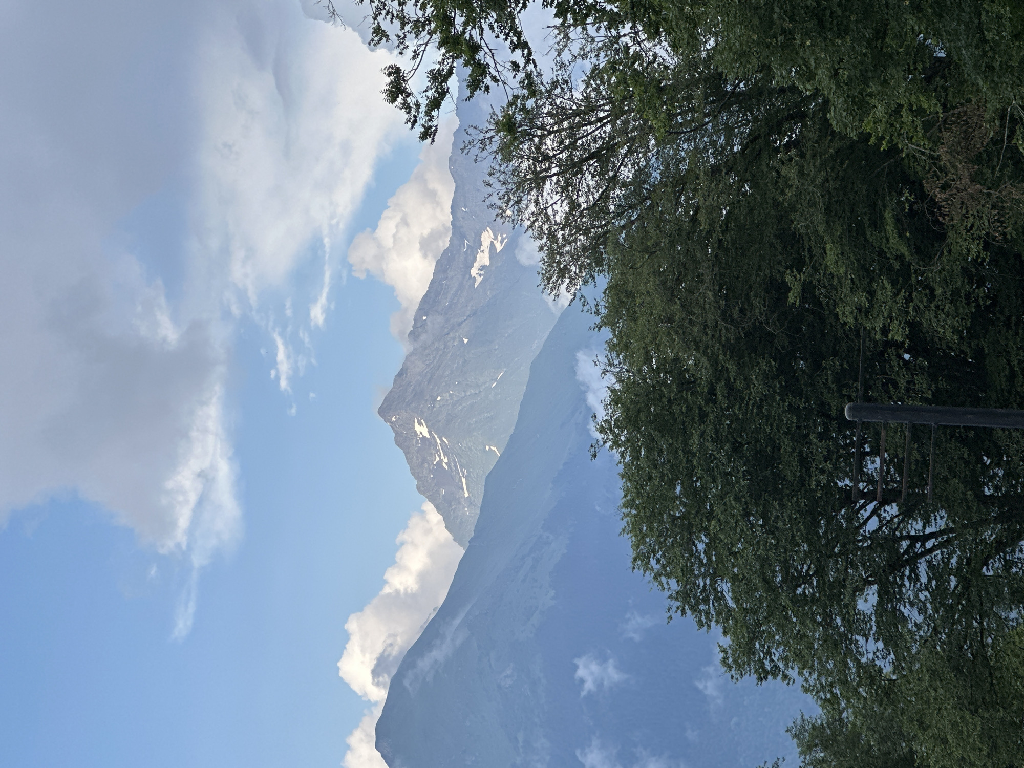 The image shows a mountain landscape with lush green trees in the foreground and majestic, snow-capped peaks in the background. The sky is a mix of blue with scattered white clouds. The scene appears to be captured in the city of Estosadok, Russia, a popular mountain resort town known for its stunning natural beauty. No people or specific actions are depicted in the image. The environment is characterized by a serene and majestic mountain vista.