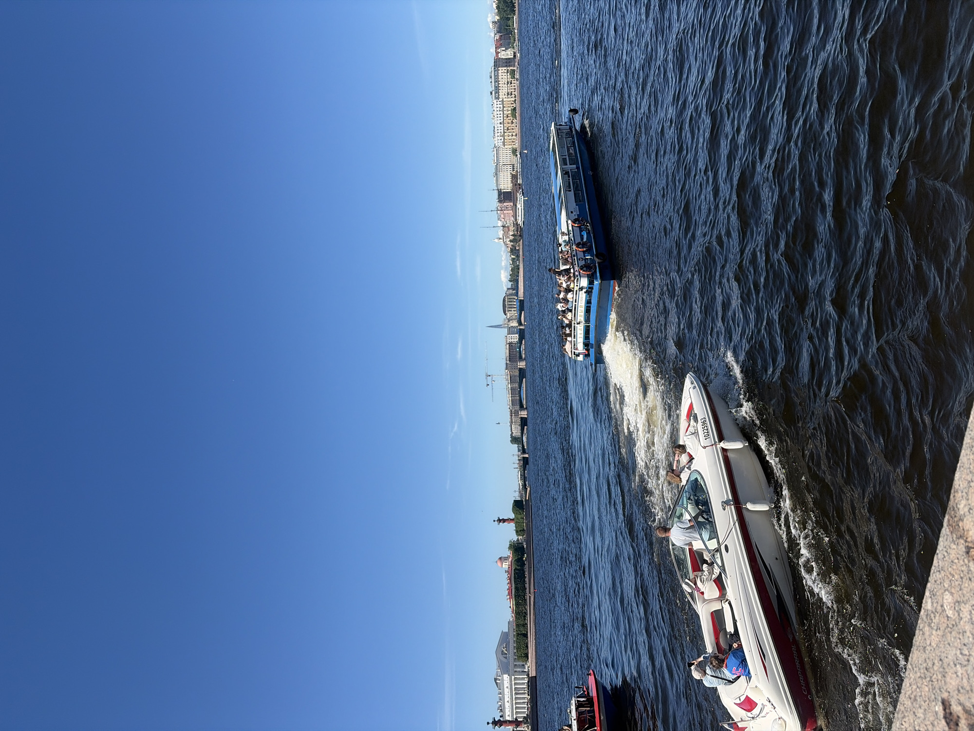 This image depicts a sunny daytime scene on the Neva River in St. Petersburg, Russia. The perspective is from an embankment, evidenced by a light-colored, textured stone parapet visible in the lower left corner. The wide river, characterized by dark blue, choppy water with ripples and waves, fills the mid-ground.
Two boats are prominently featured, both in motion and generating white wakes. In the mid-ground, towards the bottom left, a white recreational speedboat with distinctive red accents along its hull and an apparent "Chaparral" brand marking is visible. Its registration number "10225963" is displayed on the bow. Four individuals are onboard: a male is at the helm, a female passenger occupies the forward seating area, another male passenger in the bow is holding a smartphone, and a fourth person (gender indistinct) is seated towards the stern. Moving parallel to and slightly ahead of the speedboat, a larger, multi-decked blue and white passenger boat, presumably a tour vessel, traverses the river. Its upper deck is open and populated with numerous passengers. A portion of a smaller, red and blue boat is cropped on the far left edge of the frame.
The background reveals the architectural grandeur of St. Petersburg. On the left bank, the white neoclassical Old Saint Petersburg Stock Exchange building, featuring a prominent colonnade, stands on the Spit of Vasilievsky Island, flanked by two striking red Rostral Columns. Beyond these landmarks, a continuous stretch of historical buildings lines the river's edge. In the distant background, a modern skyscraper, likely the Lakhta Center, and several bridges are discernible. The sky above is clear and intensely blue, devoid of clouds.