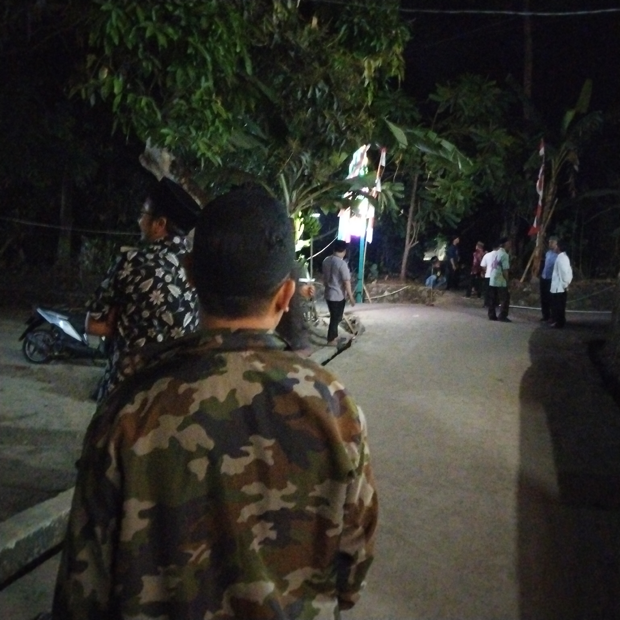 In the city of Pamulihan, Indonesia, at night, a man wearing a camouflage jacket is facing away from the camera. To his left and slightly behind him, another man is visible in profile, wearing glasses and a black shirt with a white floral pattern. A motorcycle is parked to the left of these two individuals. In the background, several people are gathered near trees, some standing and others walking along a paved area. Decorations or lights are visible in the distance. The environment appears to be an outdoor, possibly residential or communal area, illuminated by artificial light sources.