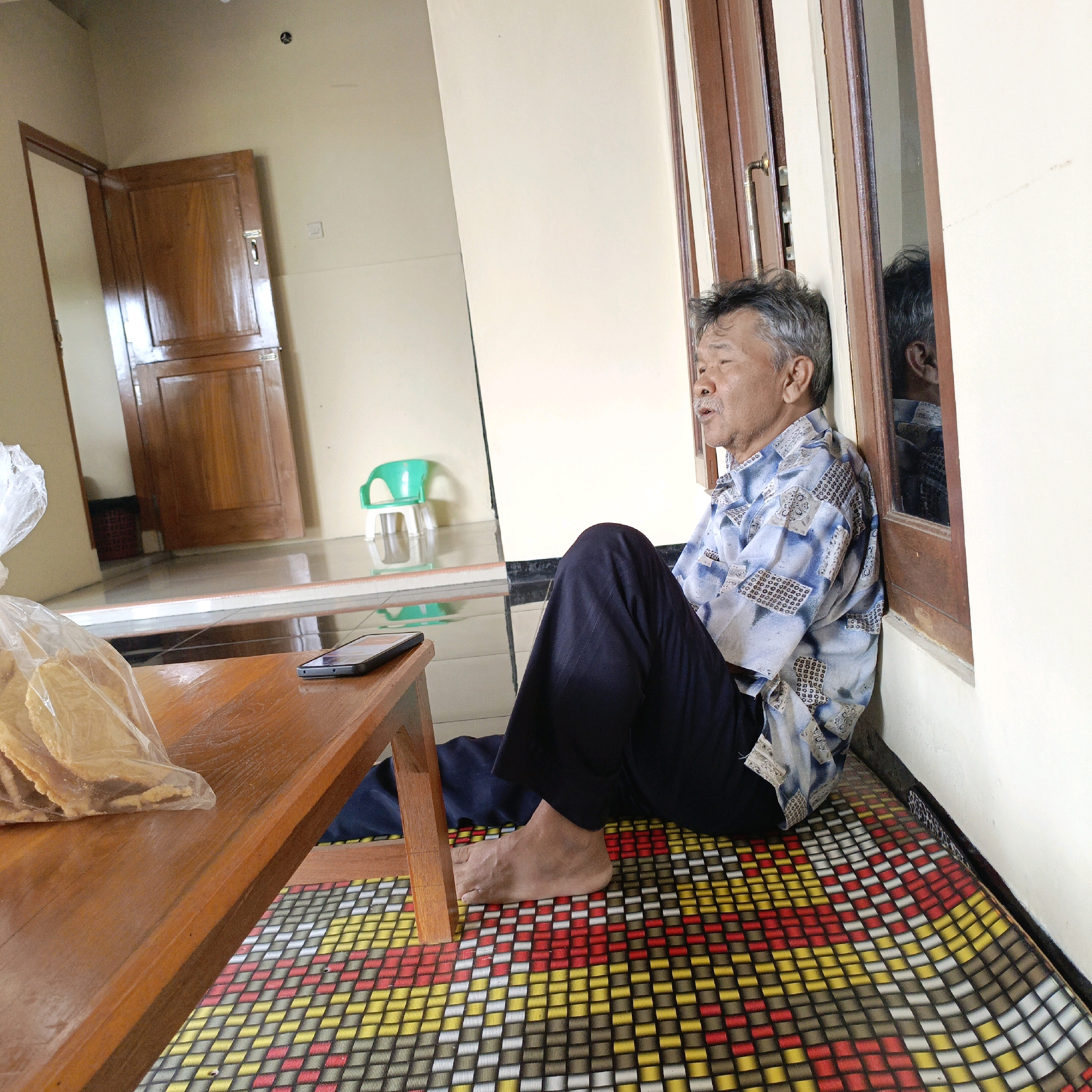 An elderly man with gray hair is sitting on the floor leaning against a wall next to a wooden door frame. He is wearing a blue and white patterned shirt and dark pants. He has his mouth slightly open as if speaking or singing. In front of him, a wooden table holds a bag of snacks and a black smartphone. The floor is covered with a colorful woven mat. To the left, a wooden door is ajar, revealing a hallway with another door. In the background, a small green plastic chair is visible. The scene appears to be inside a home in Salatiga, Indonesia, possibly a living room or entryway.