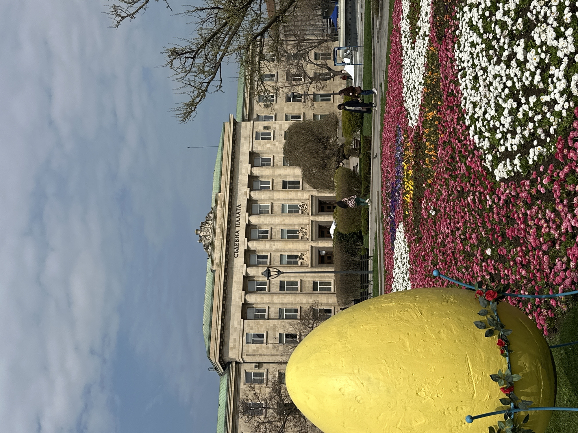 The image shows the Palace of Justice ("Съдебна палата") in Sofia, Bulgaria. The building is a large, stone structure with multiple windows and classical architectural details. In front of the building, there is a flower bed with flowers arranged in colorful rows. A large, yellow, egg-shaped sculpture decorated with roses and leaves is in the foreground. Several people can be seen in the background, near the flower bed. The sky is partly cloudy.
