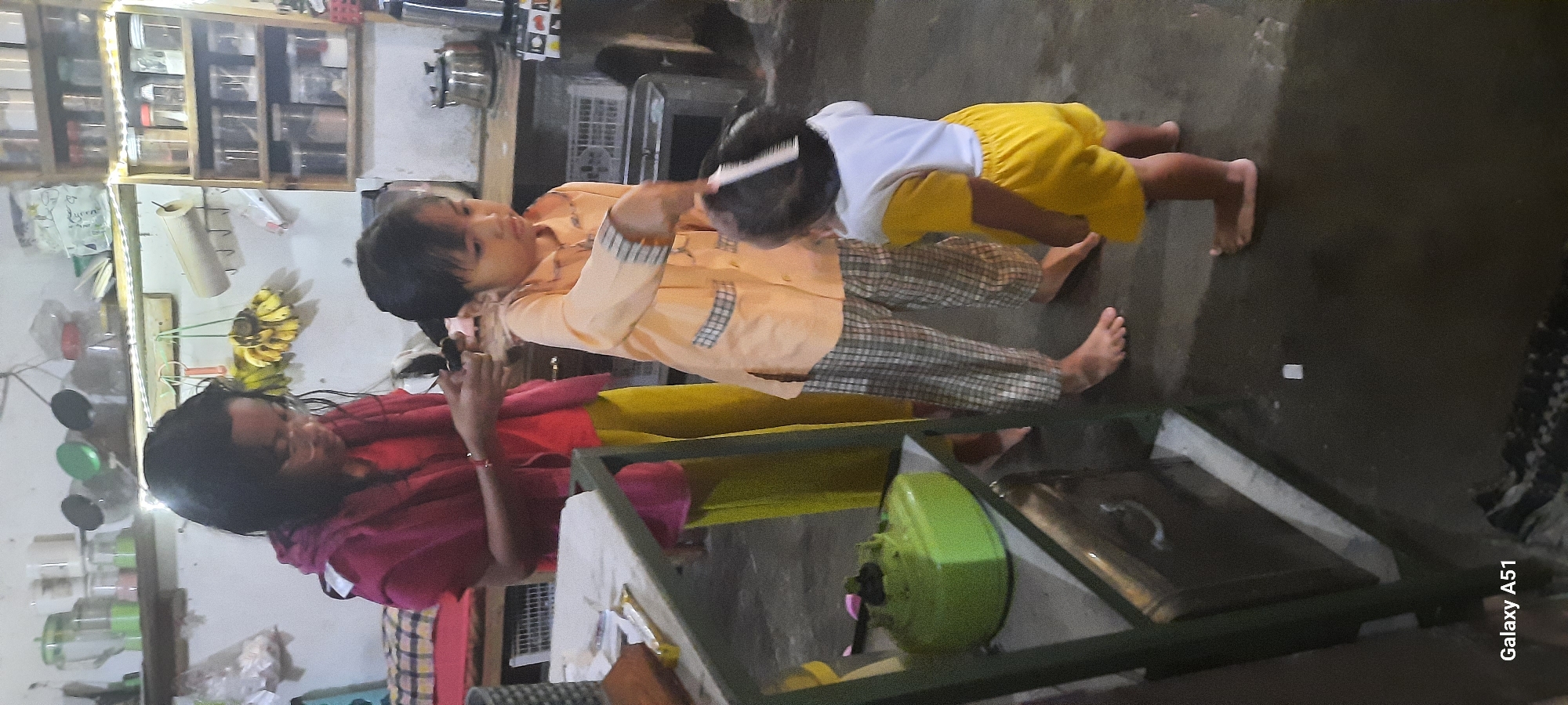 In a dimly lit interior, possibly a kitchen or workshop in Jatitengah, Indonesia, two children are engaged in an activity. A young girl with dark hair and glasses, wearing a maroon hooded top over a red t-shirt and bright yellow pants, is holding a pair of scissors and appears to be styling the hair of another child. The second child, a younger boy wearing a light orange collared shirt with plaid accents and yellow shorts, stands facing away from the camera, holding a white comb to his own hair, as if participating in the hairstyling. In the foreground to the left, a green gas stove is visible on a shelf. The environment suggests a lived-in space with shelves stocked with various items in the background, including jars and what appears to be a roll of paper towels. The floor is a dark, possibly concrete or tiled surface.