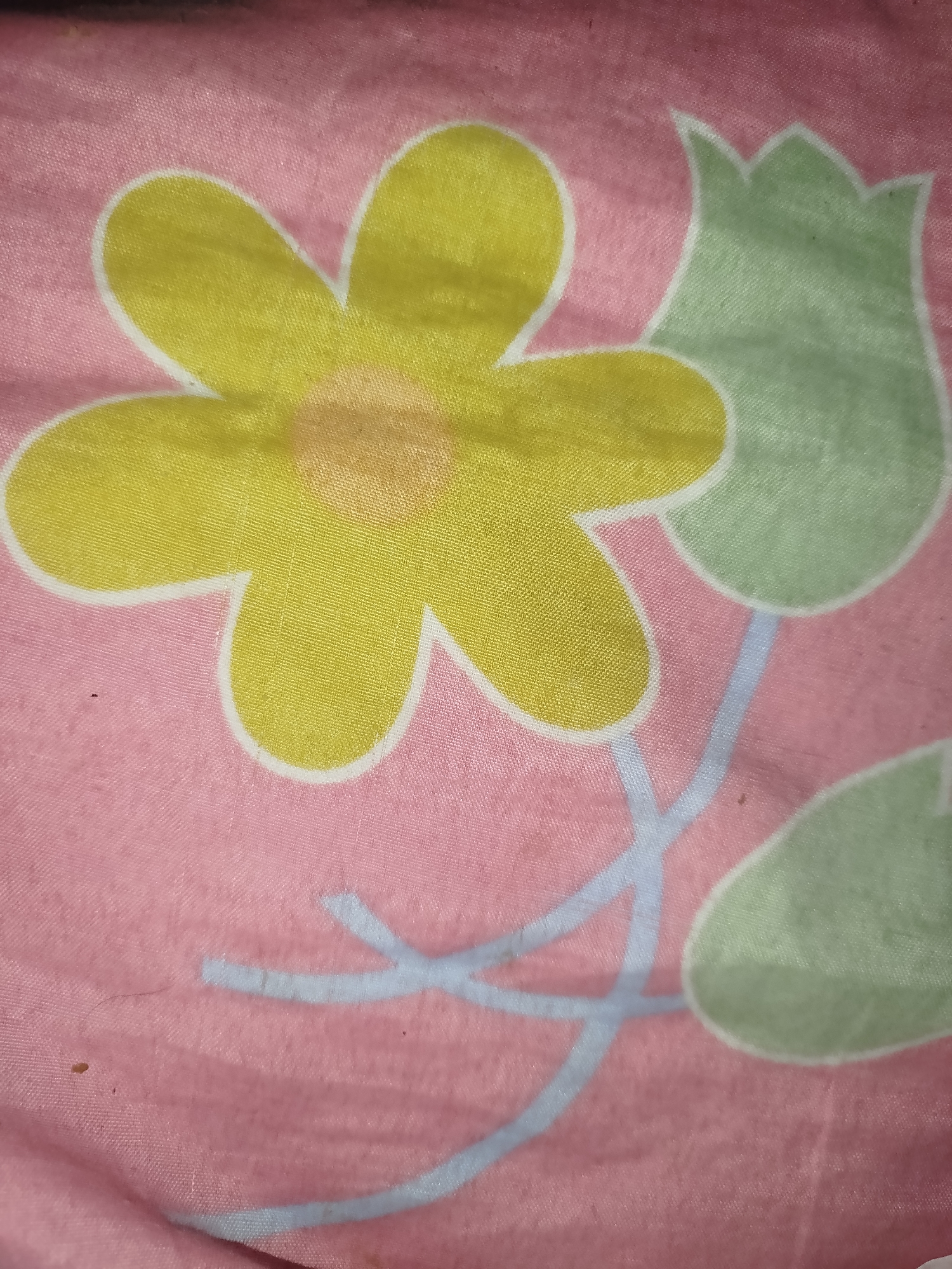 The image is a close-up, overhead shot of a pink fabric with a floral pattern. The pattern features a large yellow flower with five petals outlined in white, and two green tulip-like shapes also outlined in white. Light blue lines, representing stems and vines, connect the flowers and leaves. The fabric appears to be made of a synthetic material, possibly polyester, with a slight sheen. The lighting is diffused, and there are a few small specks and a single strand of dark hair visible on the fabric. There is no indication of the location in Lassan, Pakistan, as the image solely depicts the textile pattern.