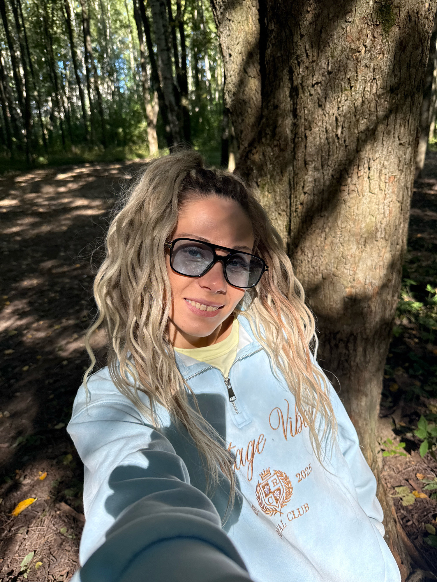 A young woman with blonde, wavy hair pulled up in a ponytail and wearing large, black-rimmed sunglasses and a light blue zip-up sweatshirt is taking a selfie in a wooded area. She is smiling at the camera. The sweatshirt has an embroidered logo with the words "Vintage Vibes" and "REGIONAL CLUB 2025". She appears to be sitting or leaning against a large tree trunk, which is in the foreground to the right. The background shows a forest with many trees and dappled sunlight on the ground. The overall scene suggests an outdoor activity or leisure time in a natural setting. The location is identified as being in the city of Dumino, Russia.