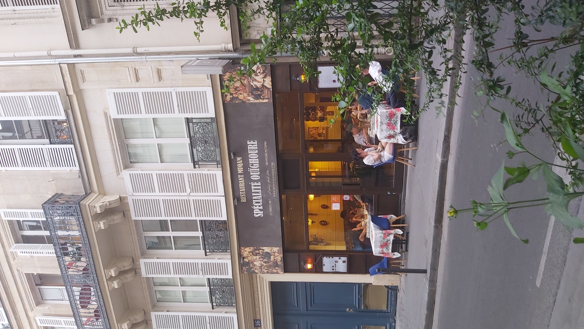 The image depicts the exterior of a restaurant named "RESTAURANT MUQAM" located in Paris, France. The restaurant specializes in "SPÉCIALITÉ OUÏGHOUR," as indicated by signage in French, Chinese, Arabic, and Russian. The establishment has a dark facade with large glass windows, allowing views into the warmly lit interior where several patrons are seated at tables. Some diners are visible inside, engaged in eating and conversation. Outside, tables with floral-patterned tablecloths are set up on the sidewalk, with blue chairs around them. A blue door is visible to the left of the restaurant entrance, and the building features traditional Parisian architecture with multiple stories, balconies, and white shutters. Lush green foliage from a nearby tree partially obscures the right side of the scene. The street in front of the restaurant is paved, with a sidewalk separating it from the building. The overall impression is a bustling street-level restaurant in a European urban setting.