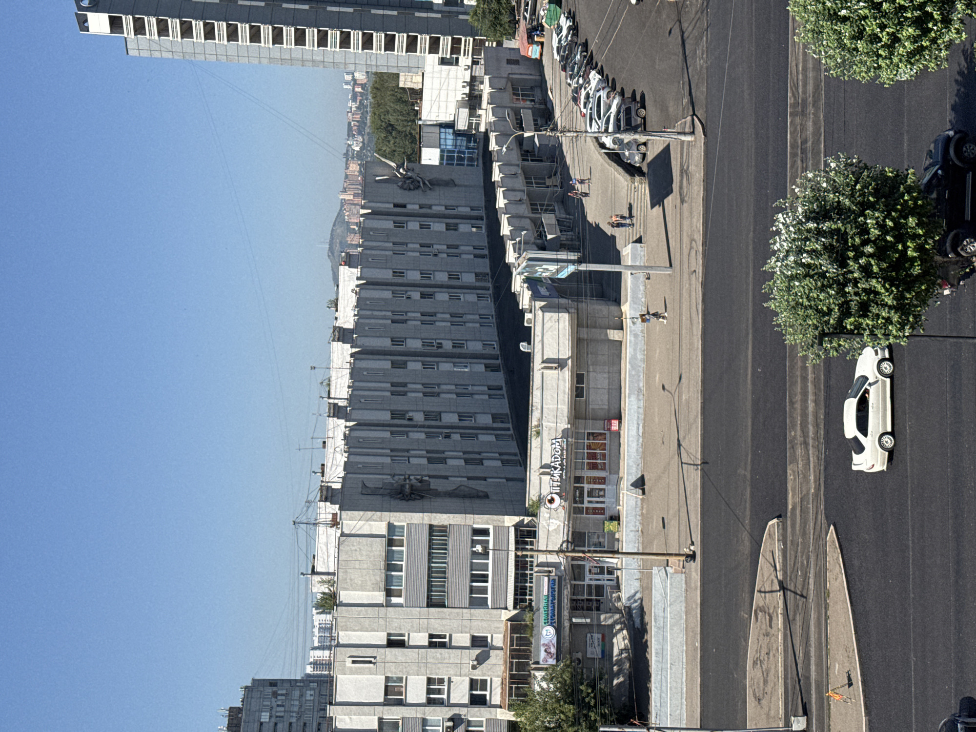 The image captures a sunny day in Krasnoyarsk, Russia, featuring a street view with several buildings and parked cars. In the foreground, a dark asphalt road is visible, with a white sedan and a black SUV parked alongside it. Two large, green trees stand on the right side of the road. Further back, a row of parked cars lines the street in front of a modern building complex with a grey facade and numerous windows. Above this, a taller, grey apartment building stands, adorned with large concrete sculptures of figures. A distant cityscape with more buildings and a hilly landscape is visible in the background under a clear blue sky. The overall scene depicts a typical urban environment on a bright, clear day.