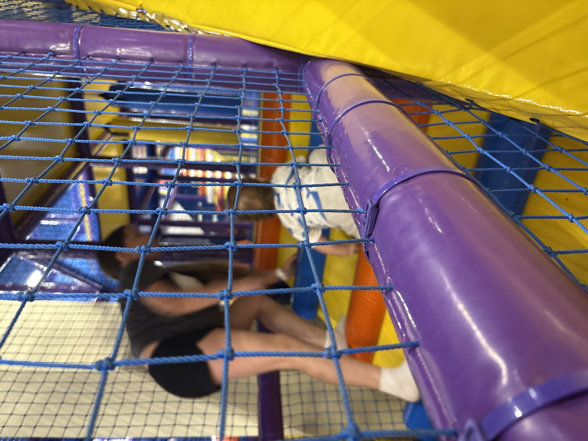 Captured from a low-angle perspective through a blue safety net, the image shows an indoor playground area, likely in a commercial space. A person in black shorts and a dark shirt is visible, prone on the floor behind the net. Another person in a white top is partially visible, reaching toward the right side of the frame. A purple padded cylindrical structure extends from the foreground to the background. The background is filled with colorful padded structures, including yellow, orange, and blue elements, which are typical of indoor playgrounds. The location is in the city of Izhevsk, Russia.