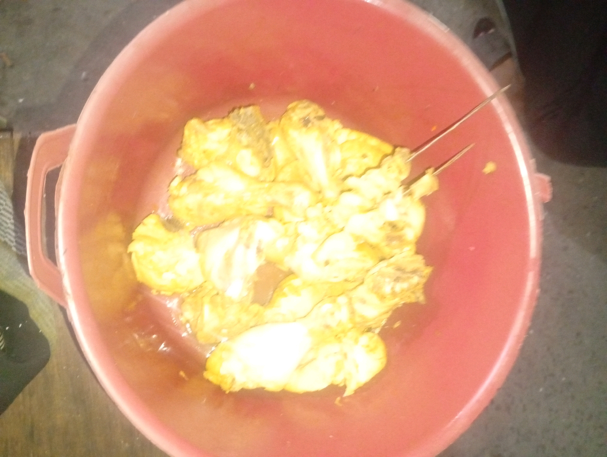 A close-up, overhead shot shows a pink bowl containing several pieces of fried chicken. The chicken appears to be seasoned and golden brown. Two metal skewers are inserted into the chicken. The bowl is placed on a dark, speckled surface. The background is blurred, but it appears to be outdoors or in a dimly lit indoor space. The image is of low resolution and out of focus, making precise details difficult to discern. It is not possible to determine the location in the city of Banowan, Indonesia from the provided image.