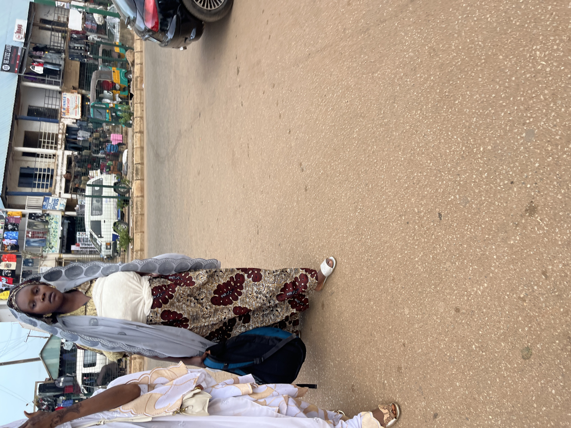 The image depicts a street scene in Bukuru, Nigeria. In the foreground, two women are walking. The woman on the right is wearing a light blue headscarf, a light-colored top, and a brown and red floral patterned skirt. She is carrying a dark blue backpack. The woman on the left is wearing a white and gold outfit and a small purse. Behind the women, there are several shops with goods displayed outside, along with parked vehicles, including a black car, a white van, and three colorful auto-rickshaws. The ground is paved with a reddish-brown asphalt. The sky is overcast.