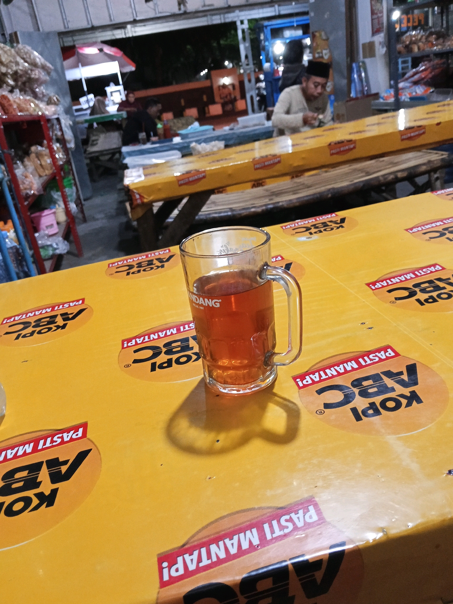 The image depicts a close-up view of a glass mug filled with amber-colored liquid, likely tea, placed on a yellow table. The table is covered with a patterned tablecloth featuring multiple circular logos with the text "KOPI ABC" and "PASTI MANTAP!". In the background, blurred figures and a dimly lit environment suggest an outdoor or semi-outdoor setting, possibly a food stall or a casual eatery. A man wearing a white long-sleeved shirt and a black peci (cap) is visible sitting at a table in the mid-ground, appearing to be eating. Shelves with packaged goods are present on the left side of the image. The overall atmosphere is casual and likely depicts a scene in Ngasem, Indonesia, given the context.