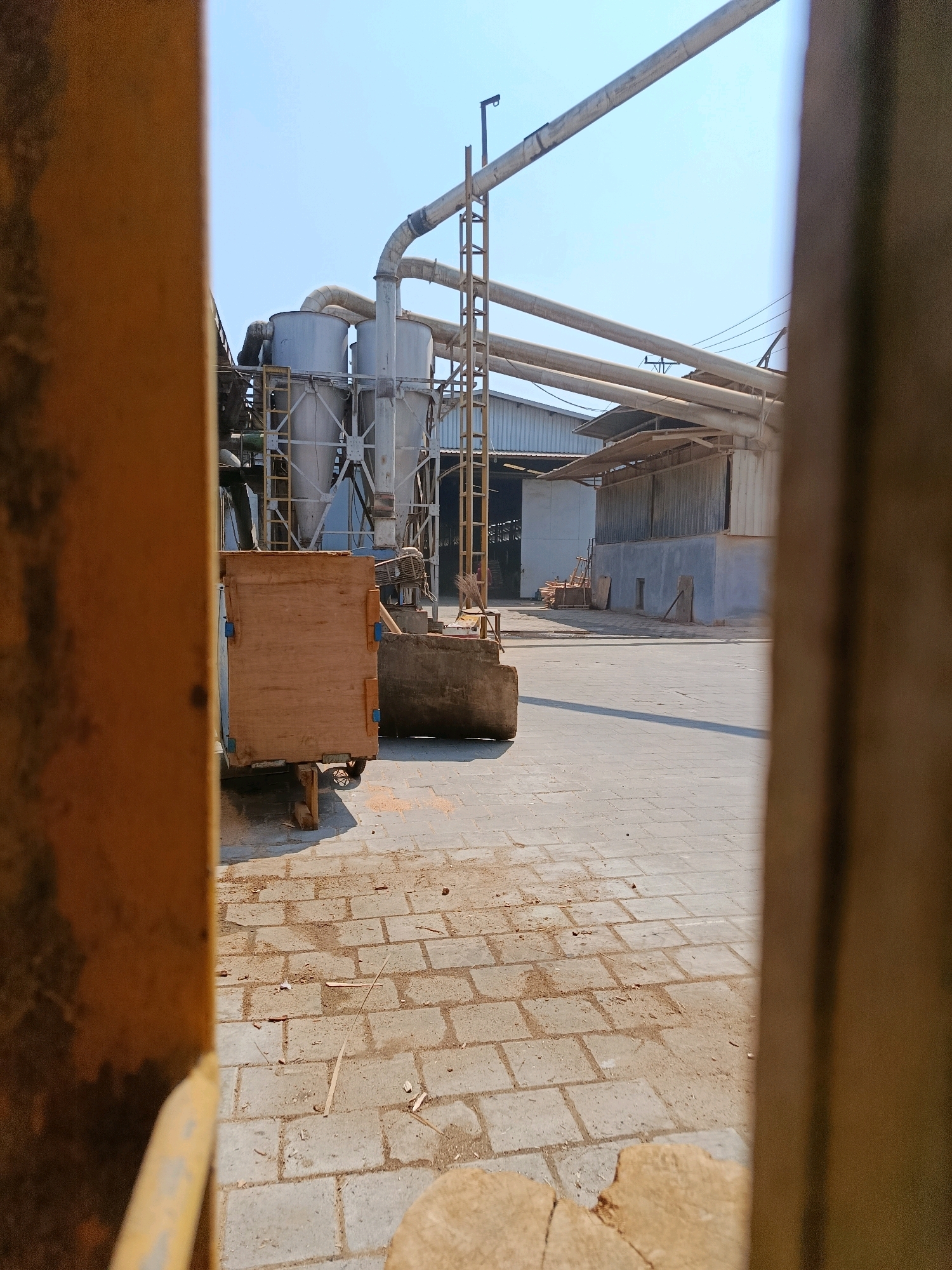 The image shows an outdoor industrial area in Kembangarum, Indonesia. In the foreground, the left side is dominated by a blurred yellow structure, possibly part of a vehicle or a gate. To the right of this, a wooden crate or box sits on a wheeled platform, partially obscuring a concrete structure and some debris on the paved ground. Beyond this, the scene opens up to a large industrial facility with several tall, conical metal silos connected by a network of pipes and scaffolding. A long, corrugated metal building with a sloped roof is visible behind the silos, and further back, another building with a white facade and a gray section. The ground is paved with rectangular interlocking bricks, with scattered wood chips and dust. The sky is clear and blue, suggesting a sunny day. There are no people visible in the image.