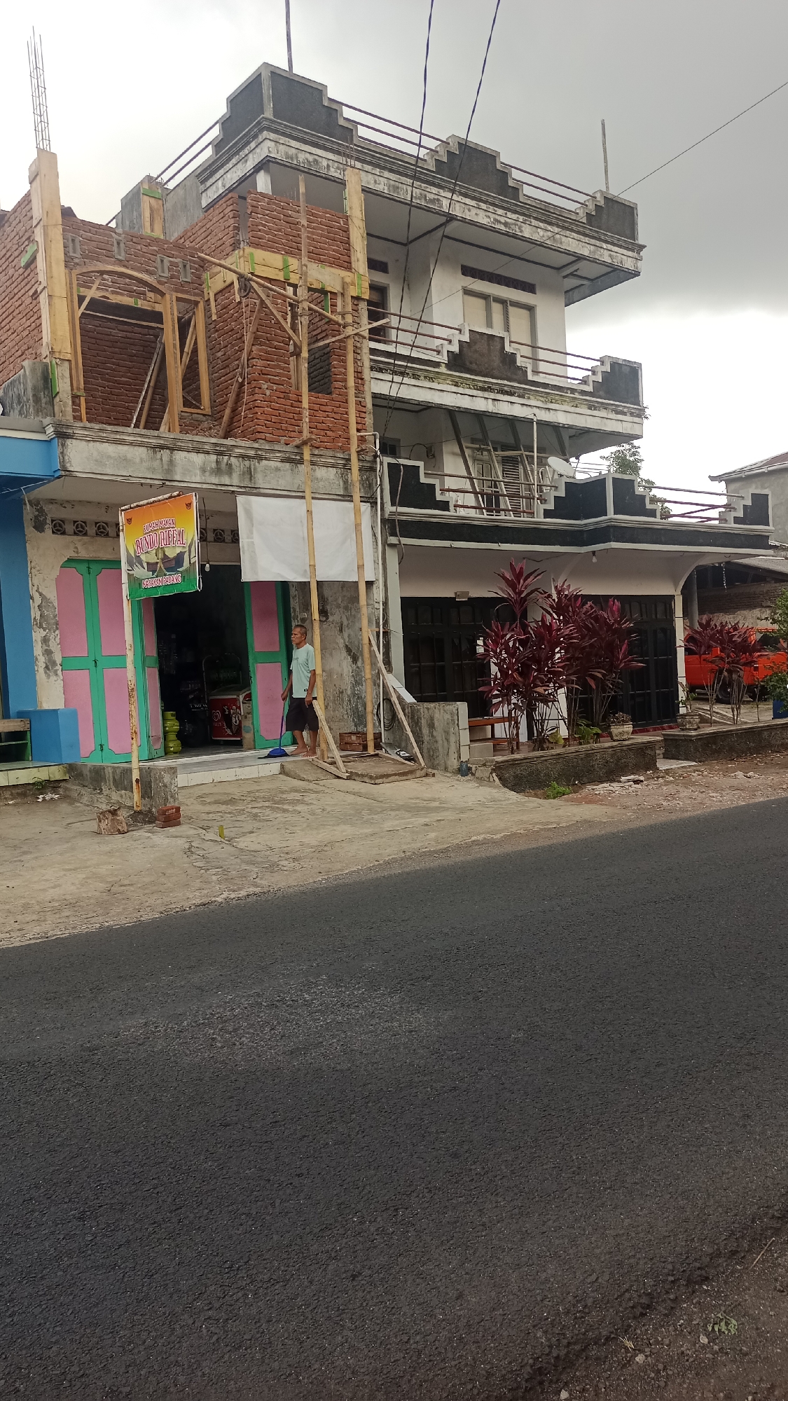 An exterior, medium-shot photograph shows a two-story building under construction and a completed adjacent building. The building on the left is partially constructed with exposed red brick and wooden scaffolding. A sign is affixed to the front of the completed building. A man wearing a light blue t-shirt and dark shorts stands outside, sweeping the pavement with a blue broom. A gas tank is visible inside the entrance of the completed building, which has pink and green doors. The completed building has a white facade with dark accents and multiple windows. Red-leafed plants are planted in planters along the front of the completed building. The street in front of the buildings is paved with asphalt. The sky is overcast. The location is the city of Karyamandala, Indonesia.